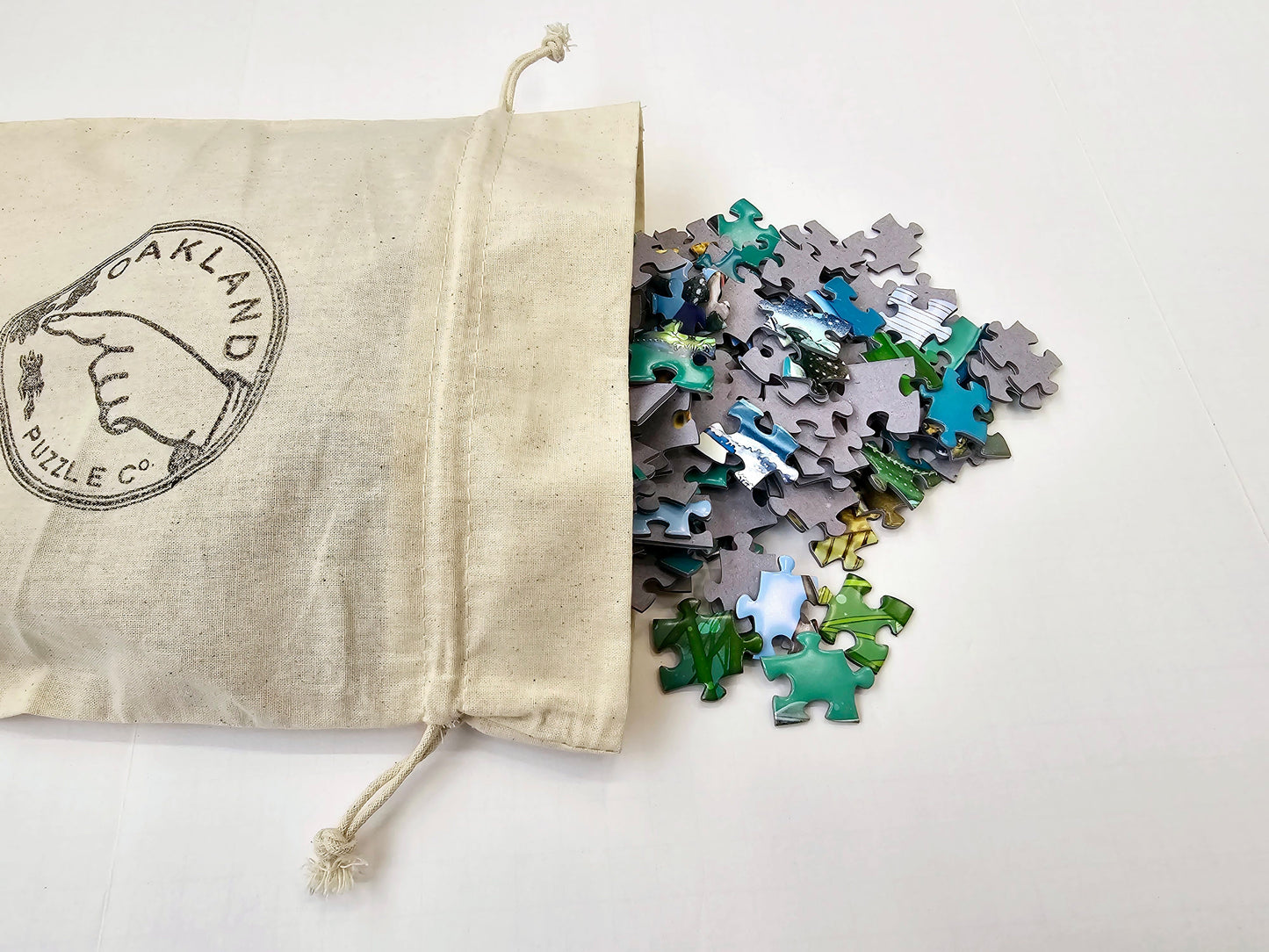 A cotton bag with the Oakland Puzzle Company logo with bright green, blue, and yellow puzzle pieces spilling out.