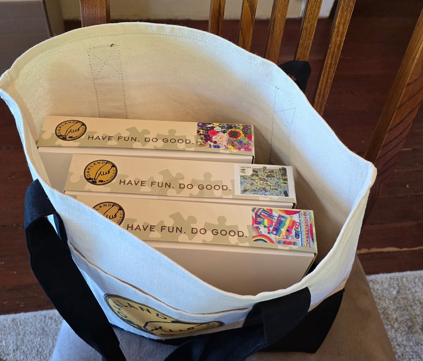 White tote bag with black straps containing puzzle boxes labeled 'Have Fun. Do Good.' on a wooden floor.
