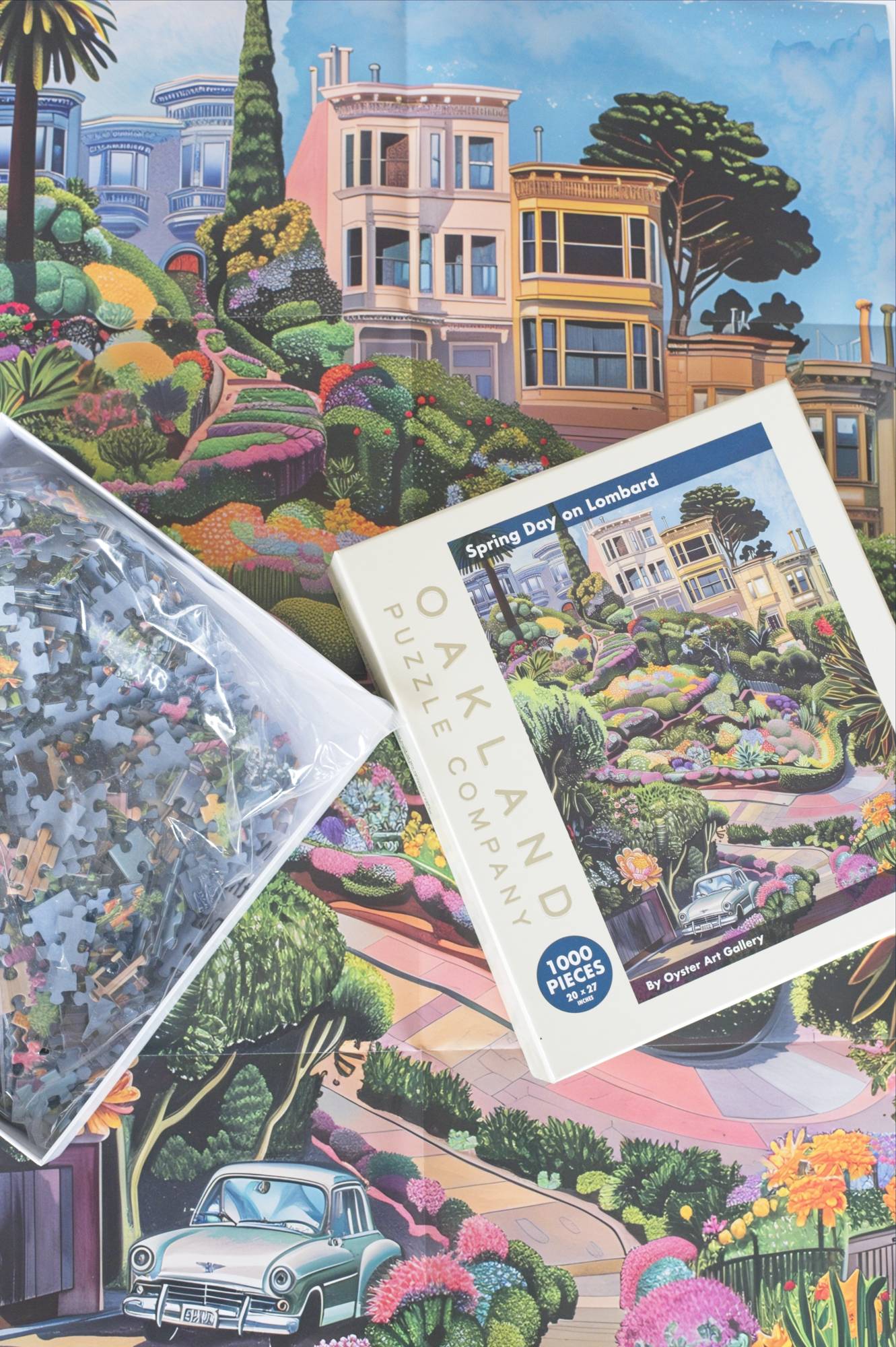 A photo of a large poster laying flat with an open box displaying a resealable plastic bag filled with colorful puzzle pieces and a lid showing a whimsical image of Lombard Street.