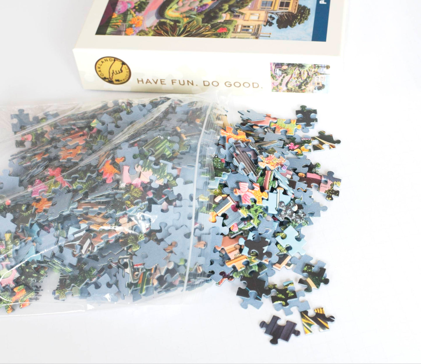 A photo of colorful puzzle pieces with blue board spilling out of a resealable plastic bag with a puzzle product box in the background labeled Have Fun. Do Good.