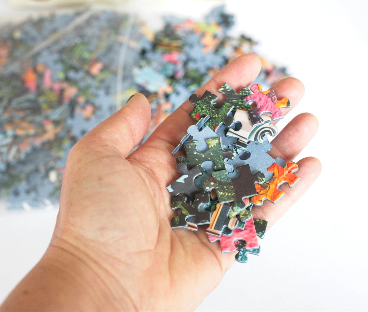 A hand holding a variety of traditionally shaped colorful puzzle pieces with a resealable plastic bag spilling pieces out of focus in the background.