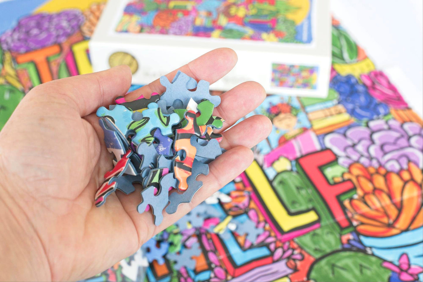 A hand holds many small oddly shaped puzzle pieces made with blue board with a large poster and puzzle box out of focus in the background.