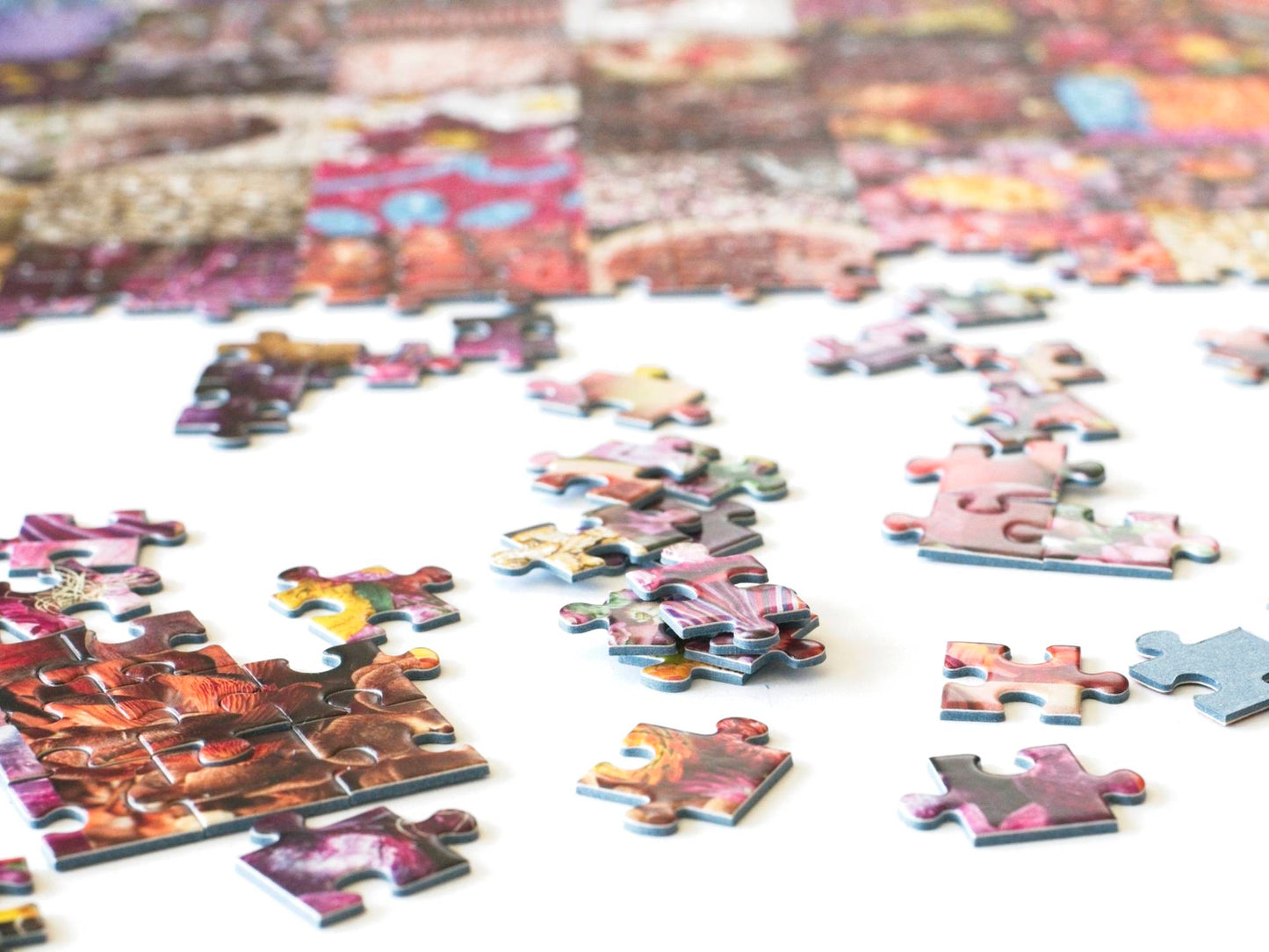 A stack of puzzle pieces with a few partially completed sections, and other pieces scattered around with a large section of completed puzzle all mostly various shades of red.