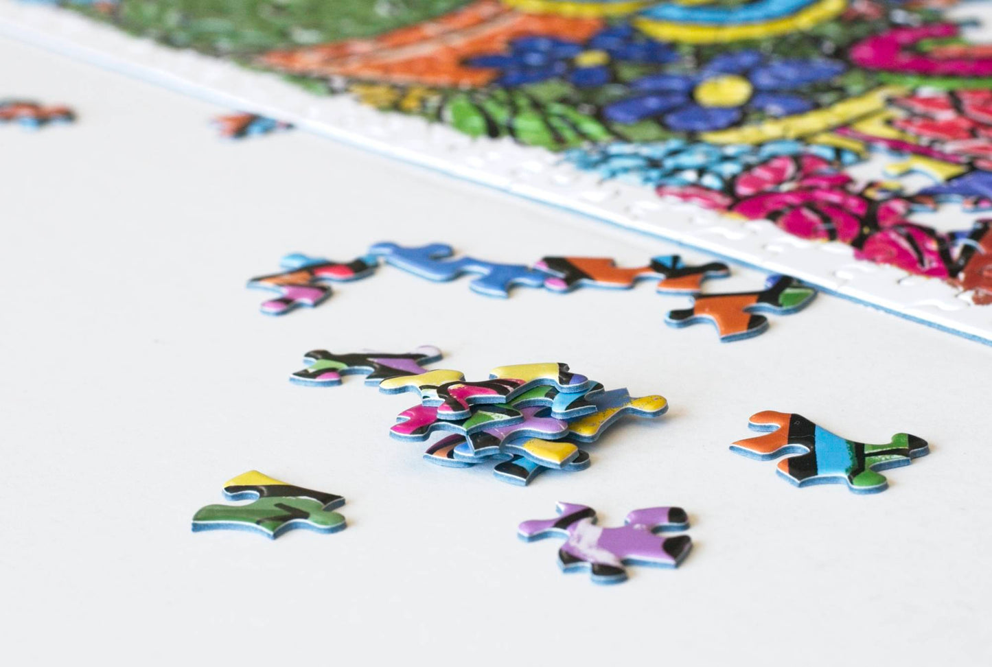 Brightly colored stacked jigsaw puzzle pieces with a few scattered pieces surrounding them with a nearly complete jigsaw puzzle out of focus in the background.