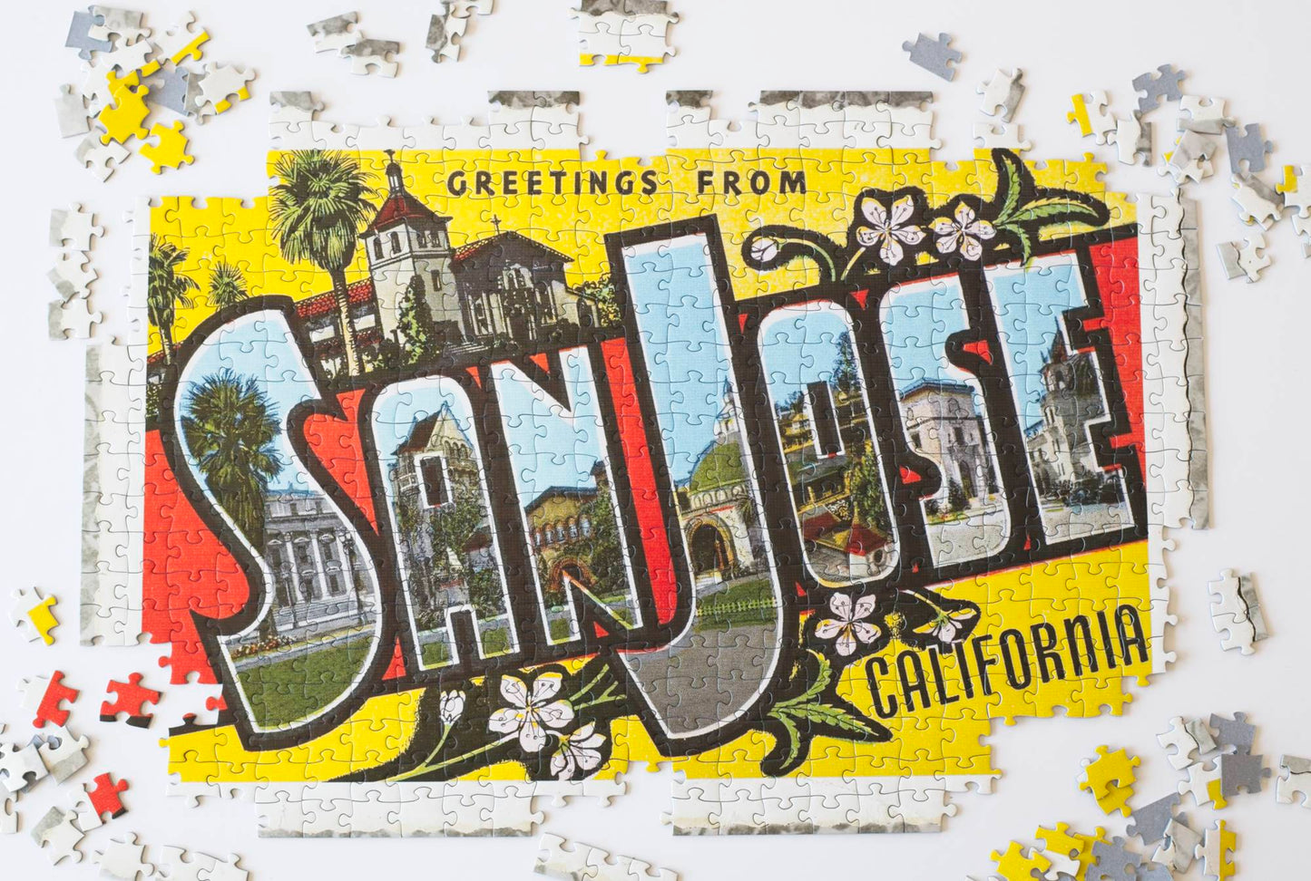A partially completed jigsaw puzzle depicting a vintage postcard with block letters that read Greetings from San Jose California with frayed edges laying on an old cloth. Buildings are featured in the letters of the words San Jose with pieces scattered around it.