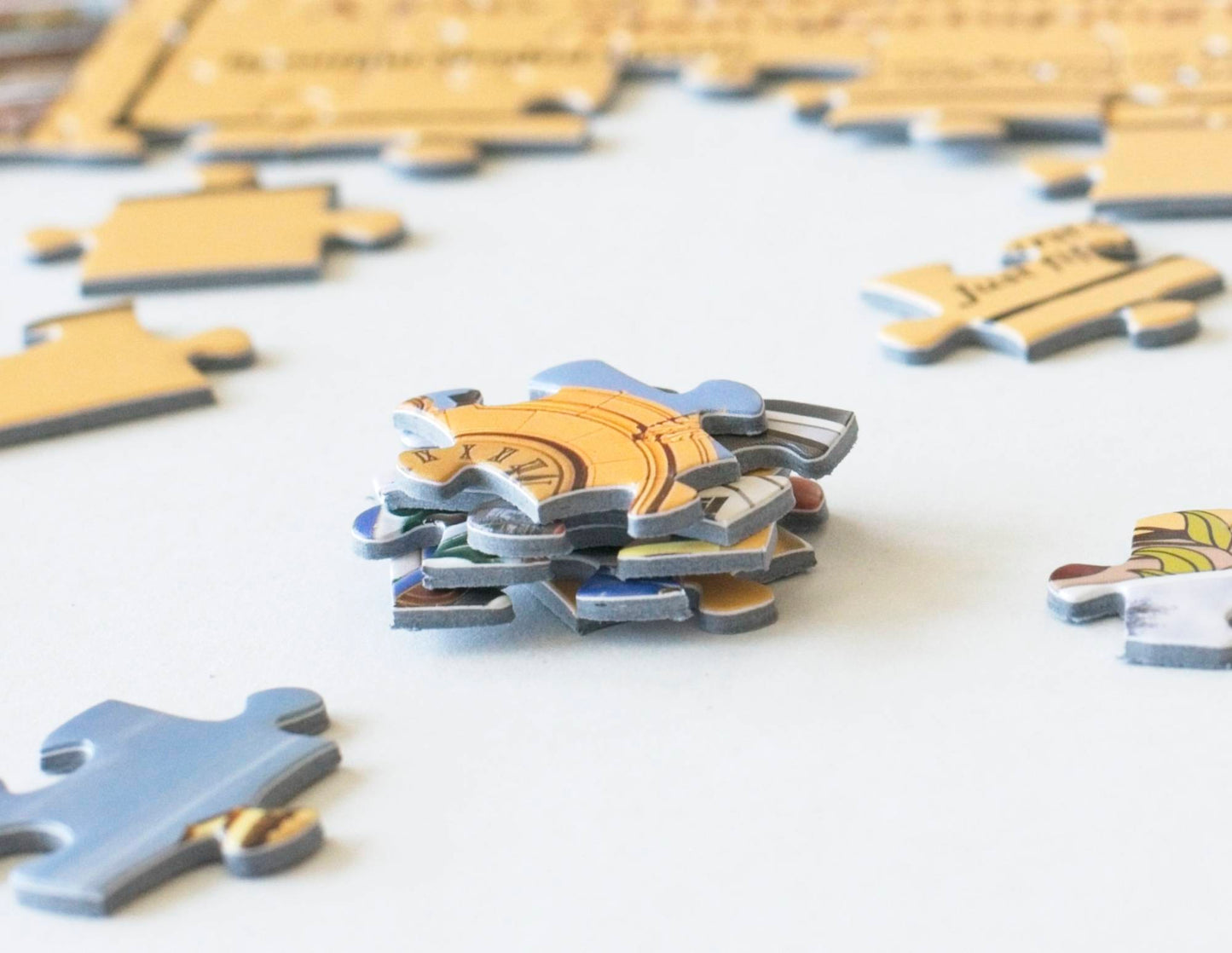 A stack of sturdy and colorful jigsaw puzzle pieces. with a complete jigsaw puzzle section in the background and surrounded by scattered pieces.
