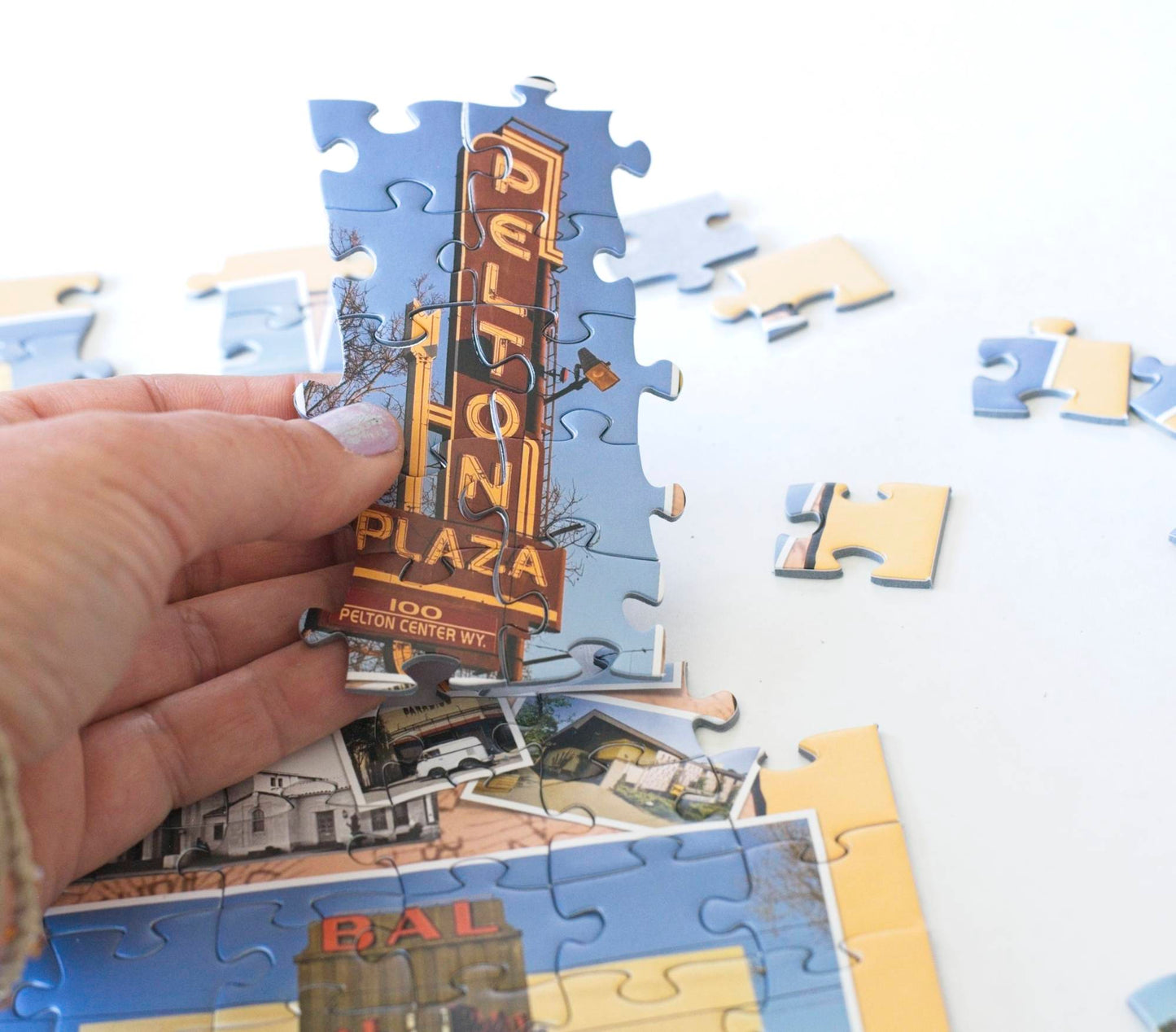 A close up of a hand  holding a section of completed jigsaw puzzle pieces  showing a Pelton Plaza sign near an incomplete section of the puzzle as if about to place the pieces.