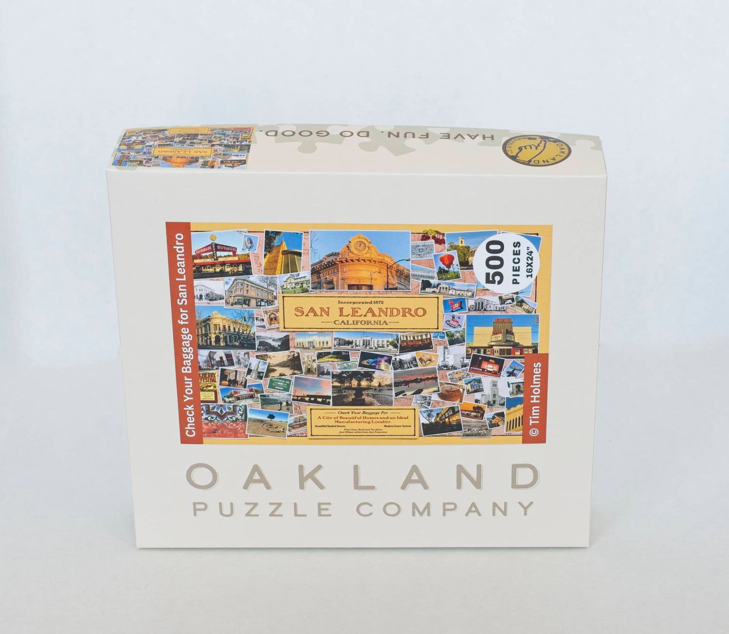 A photo of a product box branded Oakland Puzzle Company, labeled with an image of the puzzle front with the title "Check Your Baggage for San Leandro Copyright Tim Holmes.