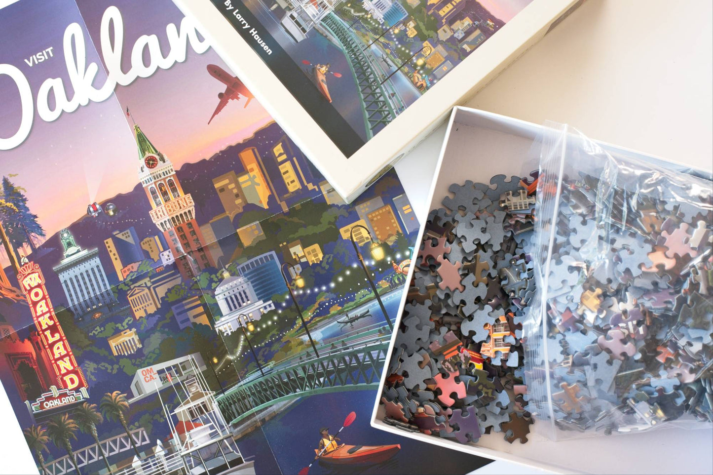 The reference image for a puzzle with a cityscape design along with an open box filled with blue backed puzzle pieces spilling out of a resealable plastic bag.