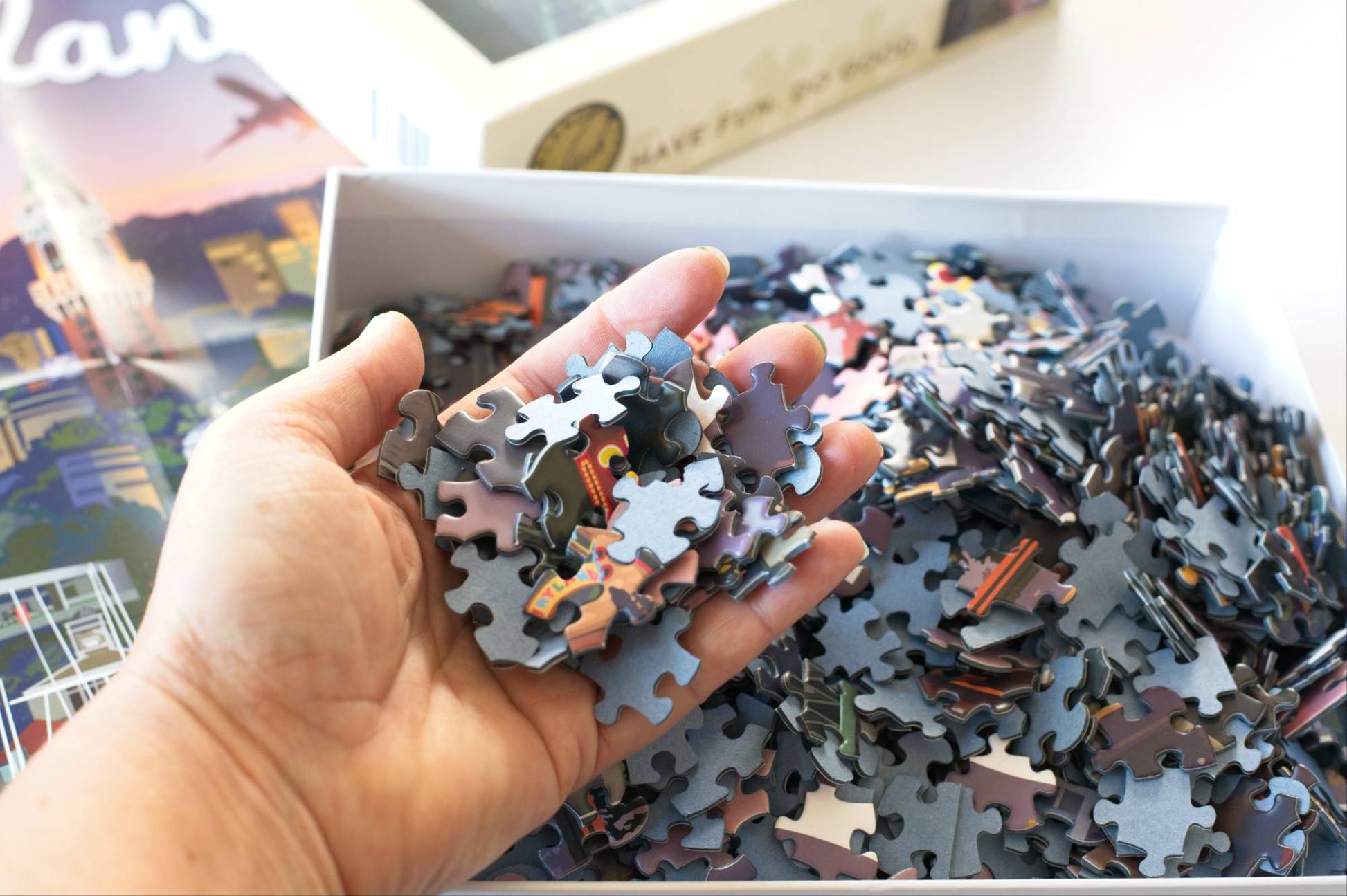 Hand holding puzzle pieces with an open box of puzzle pieces in the background