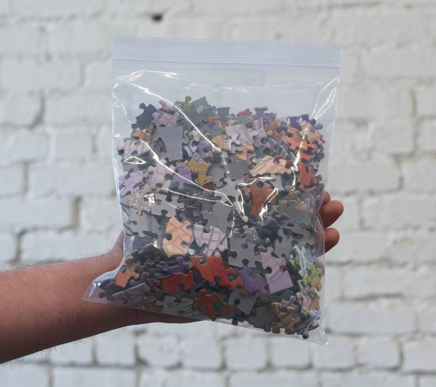Hand holding a clear plastic bag filled with assorted puzzle pieces against a light brick wall.