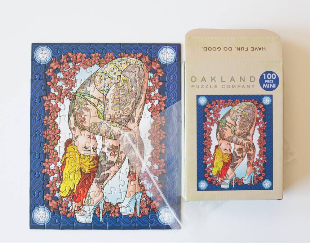 A completed 100-piece jigsaw puzzle and its matching box from Oakland Puzzle Company with a resealable plastic bag featuring an Art Deco style illustration of a woman tattooed woman in high heels surrounded by red flowers.