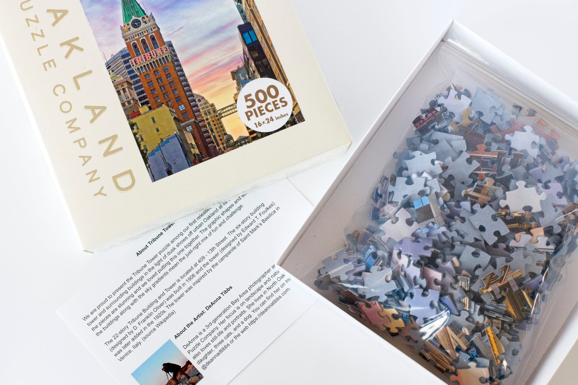 An open puzzle box with a puzzle guide. The box top is branded Oakland Puzzle Company and features the Tribune Tower image on the front. The box bottom has a plastic bag filled with puzzle pieces.