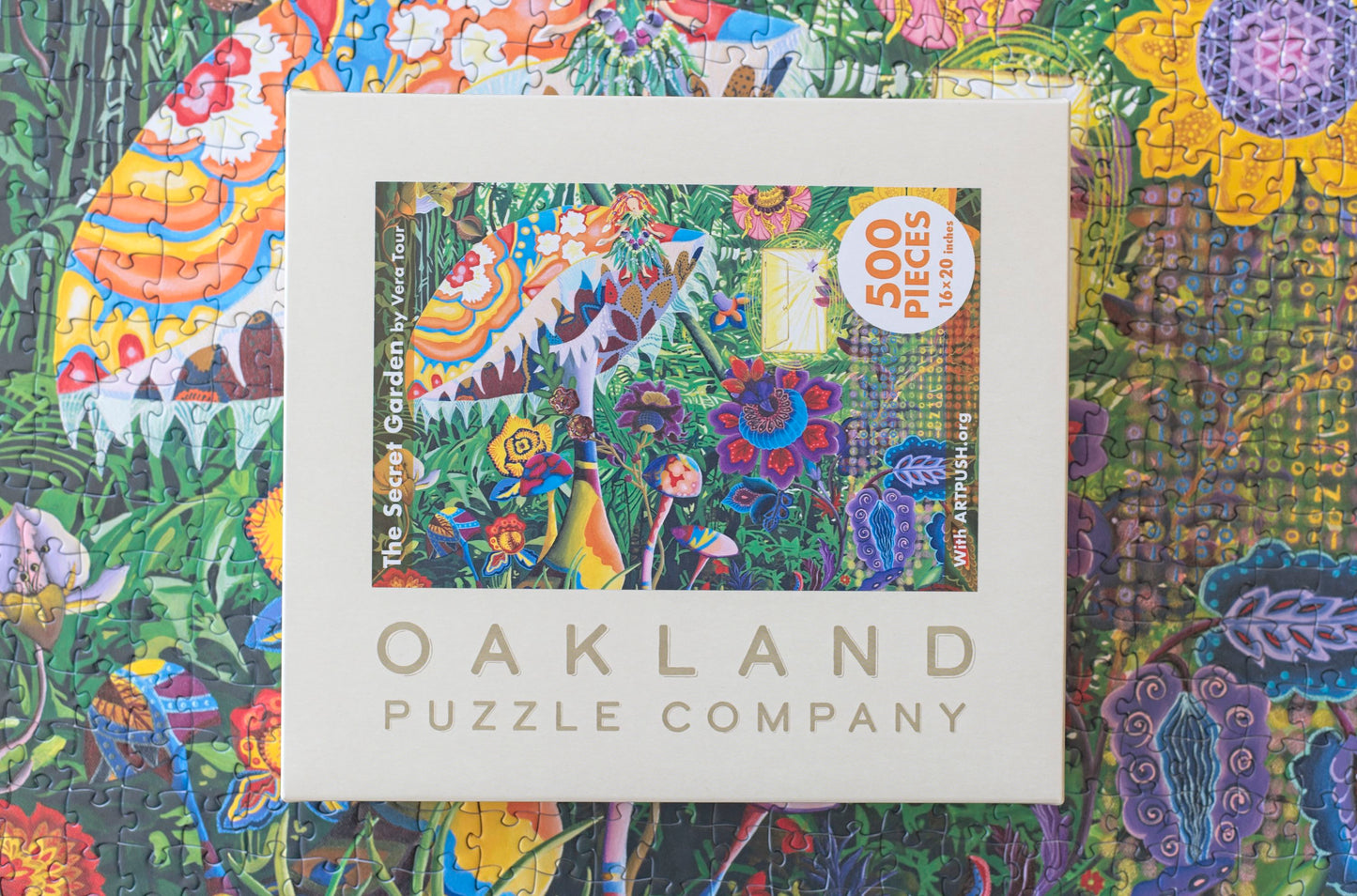 A completed jigsaw puzzle with the box for the puzzle sitting on top branded Oakland puzzle company. The puzzle has a psychadelic cene with mushrooms and flowers