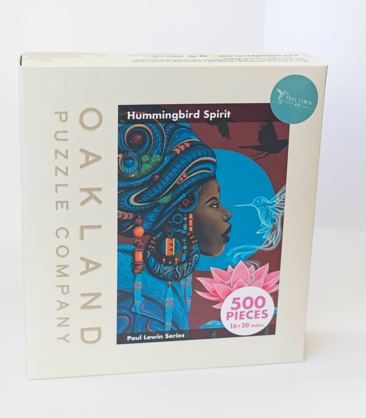 a box branded Oakland Puzzle Company with a painted image of an Afrofuturist styled woman adorned with with, flowers, beads, and feathers, and surrounded by birds. Labeled Hummingbird Spirit by Paul Lewin