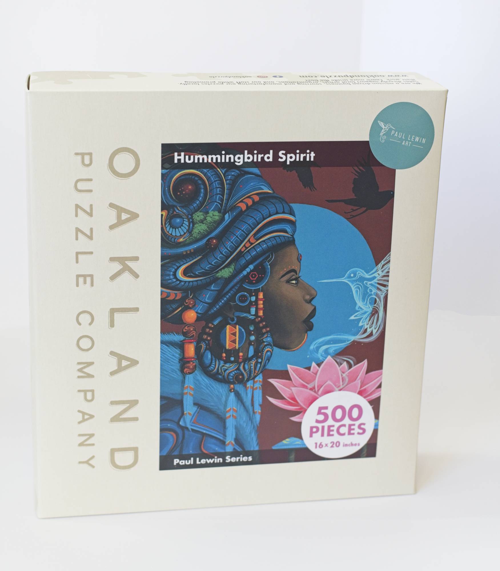 a box branded Oakland Puzzle Company with a painted image of an Afrofuturist styled woman adorned with with, flowers, beads, and feathers, and surrounded by birds. Labeled Hummingbird Spirit by Paul Lewin