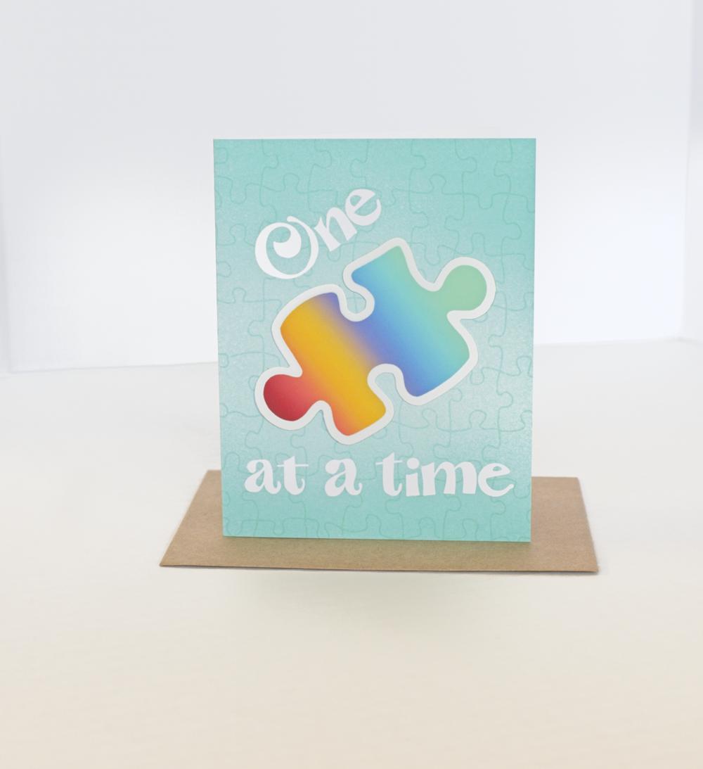 A greeting card with a jigsaw puzzle background and a rainbow sticker shaped like a puzzle piece. Text on the card says One 🧩 at a time 