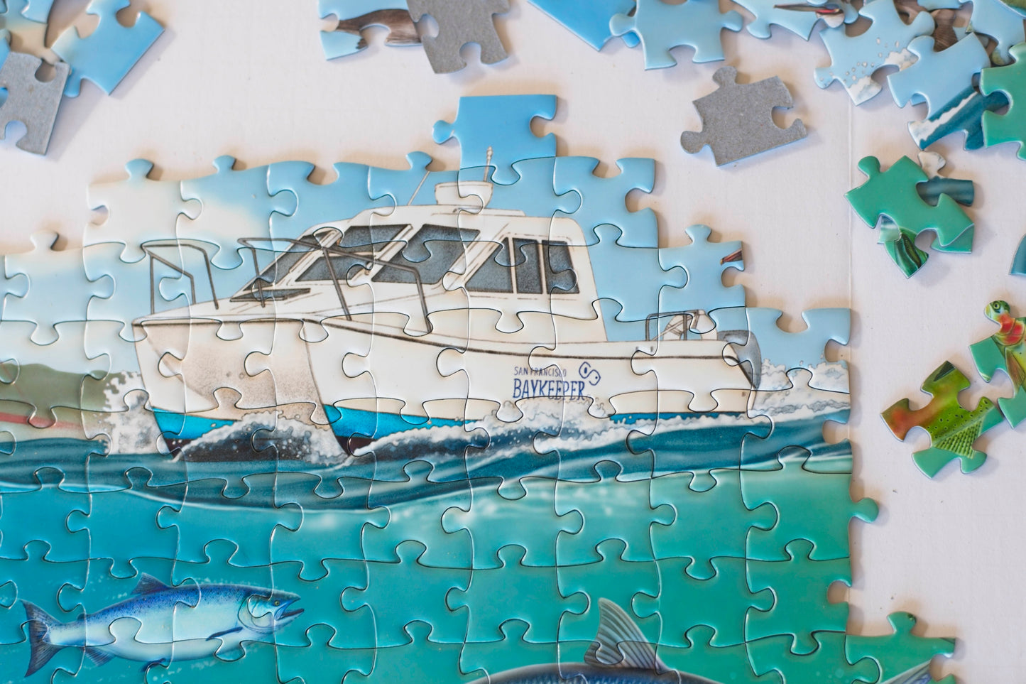 A completed section of jigsaw puzzle showing a boat marked San Francisco Baykeeper on the water with fish below. Puzzle pieces area scattered above.