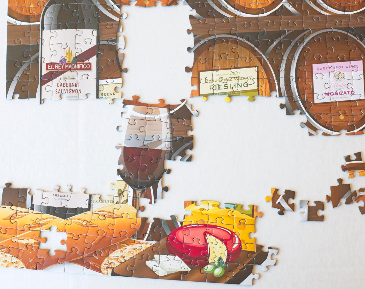 Two areas of a partially completed jigsaw puzzle that look like they will join together. One features a cheese board and the other has wine barrels and a bottle.