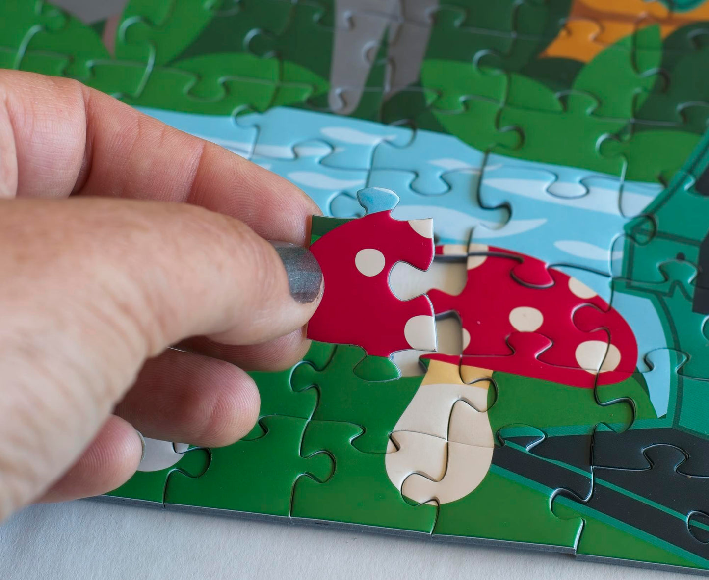 A hand placing a puzzle piece of a red mushroom cap with white spots onto a completed area of a colorful puzzle