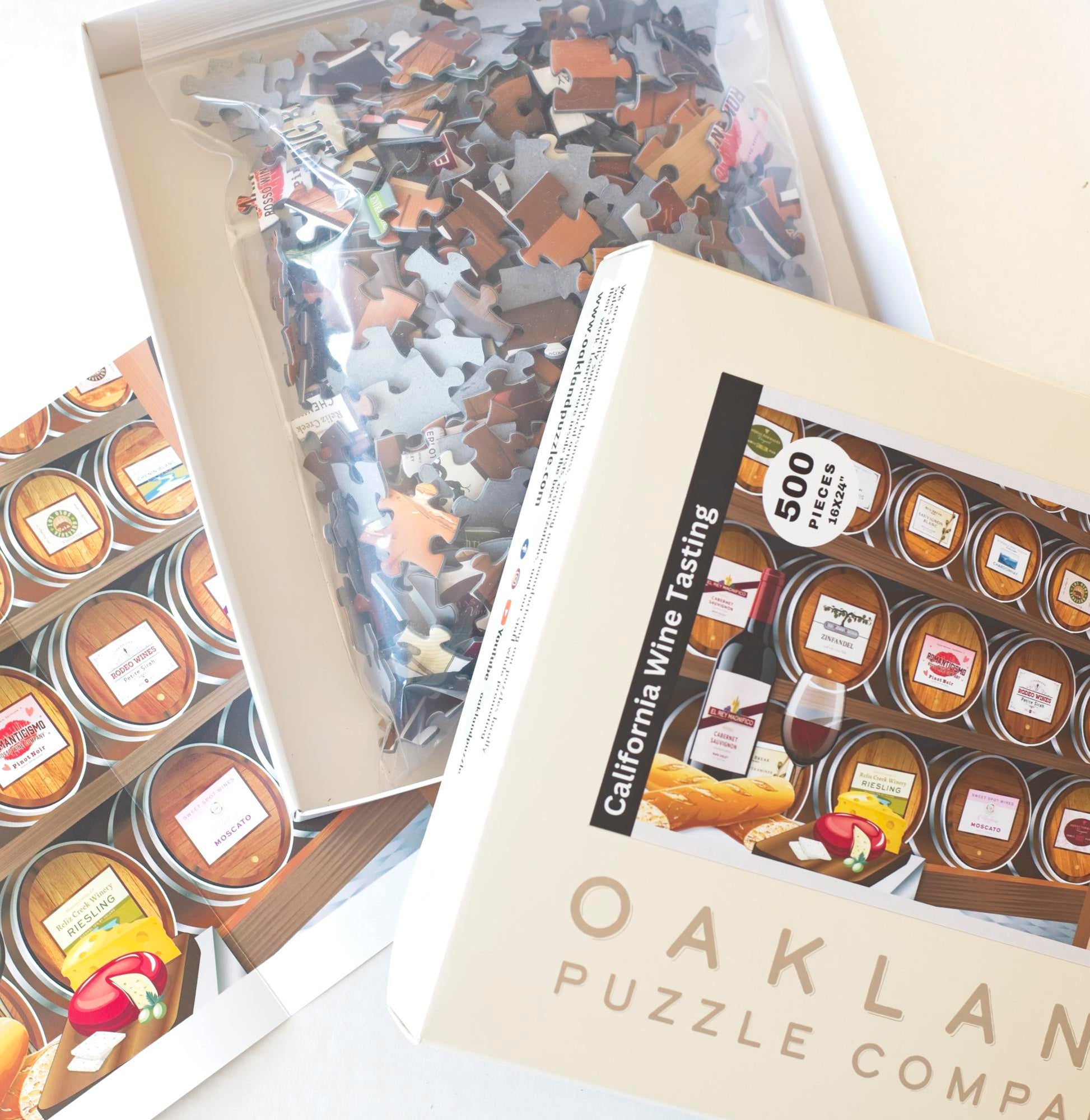 A close up of an opened box of the Oakland Puzzle Company California Wine Tasting jigsaw puzzle. Inside the box is a resealable back with jigsaw puzzle pieces and behind the box is a puzzle poster.
