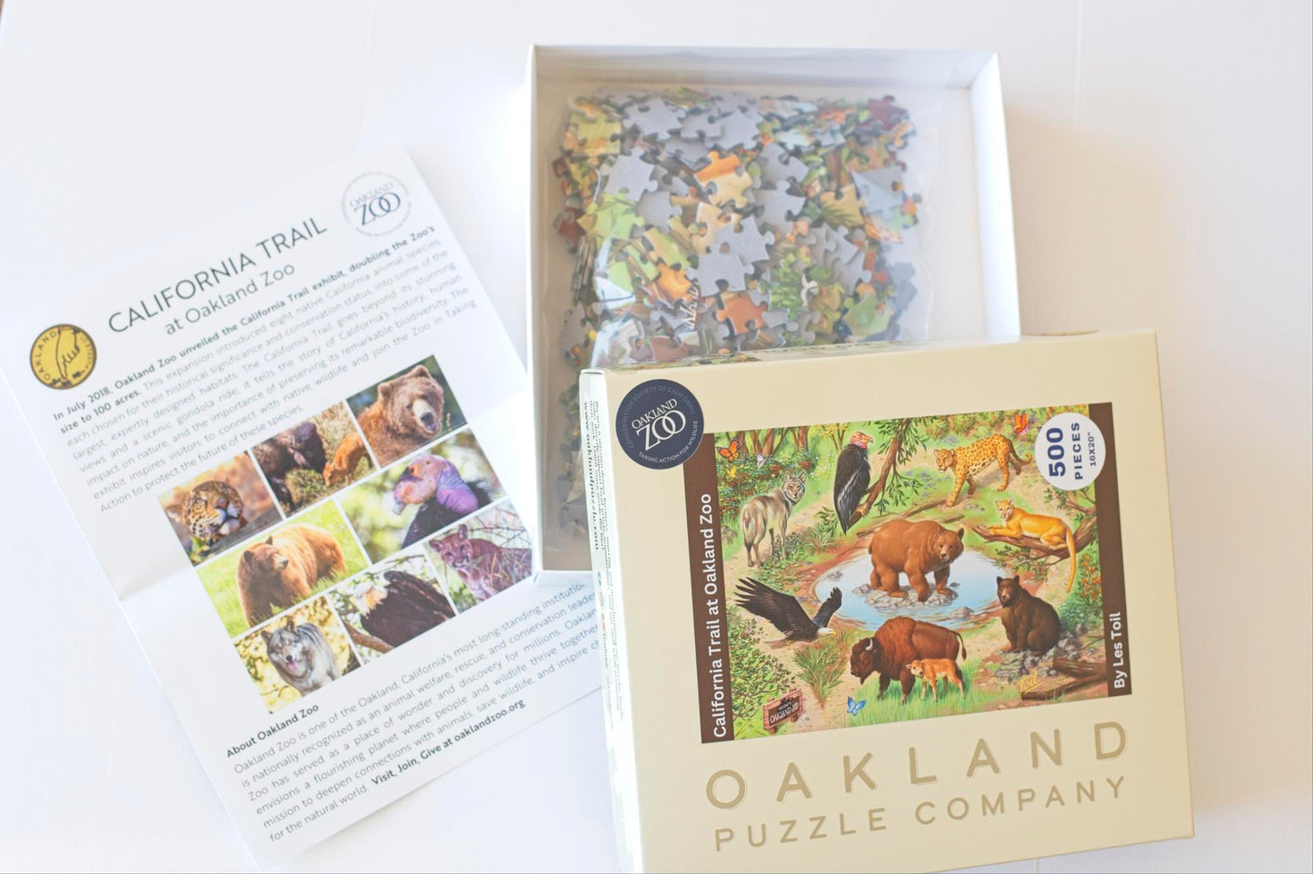 An open box branded Oakland Puzzle Company featuring the California Trail at the Oakland Zoo puzzle. The box bottom has puzzle pieces and next to it is a guide to the puzzle with information about the zoo.