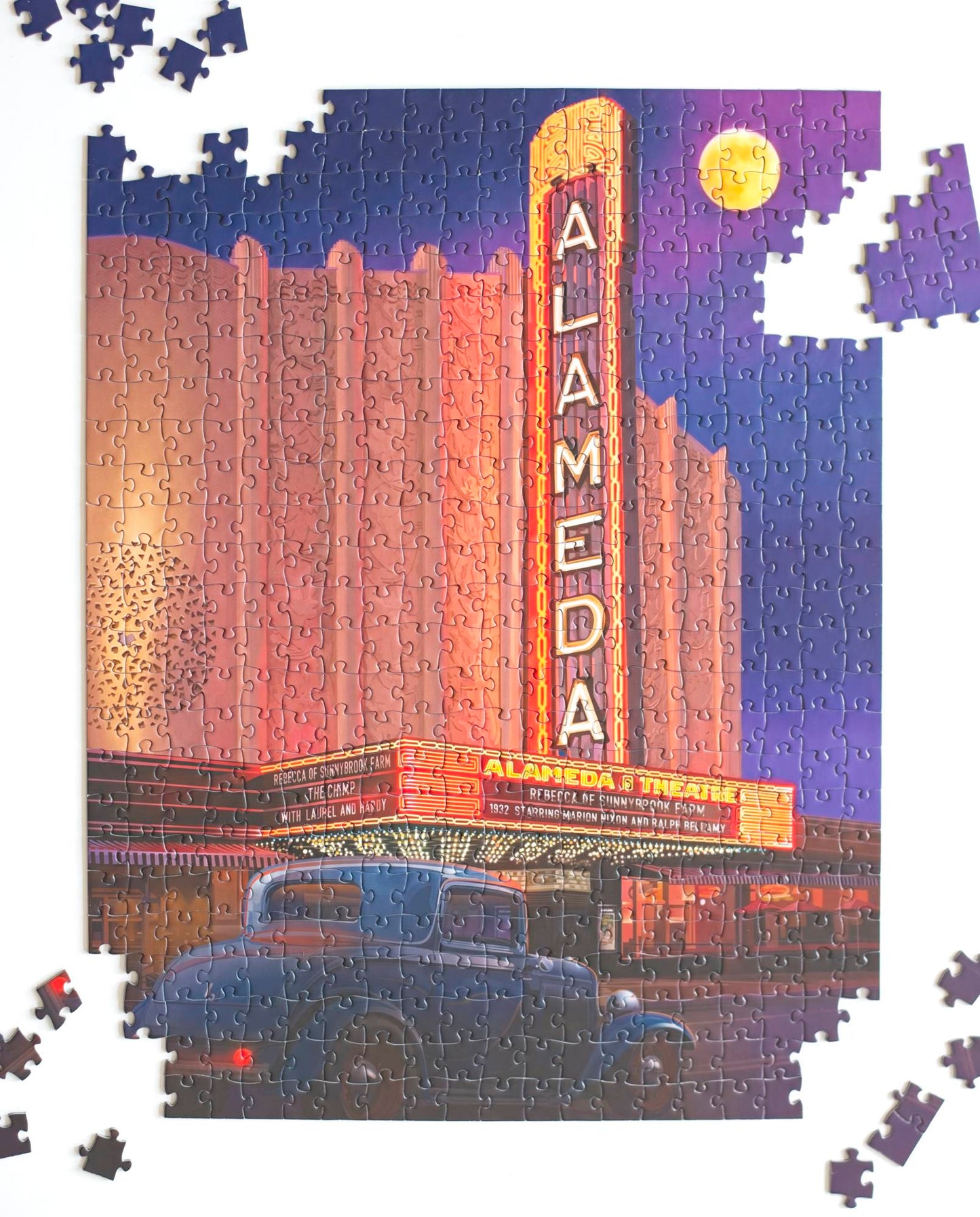 A nearly completed jigsaw puzzle featuring the art deco Alameda Theater at night with marquee lit and a classic car in front. Several sections and pieces are scattered around the outside of the puzzle.