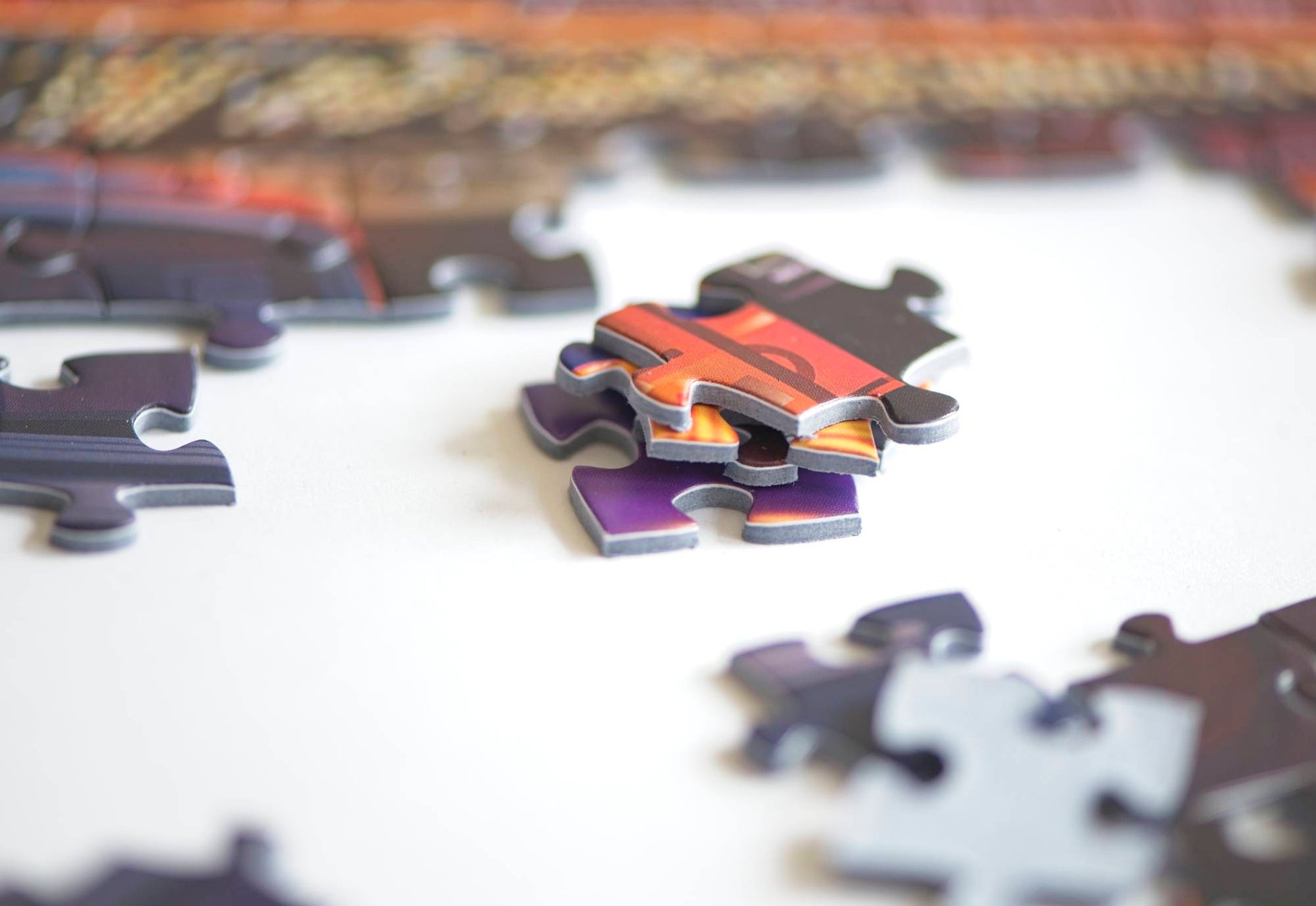 A close up shot of a stack of thick and colorful jigsaw puzzle pieces on blue board surrounded by out of focus completed sections of the puzzle and scattered pieces.
