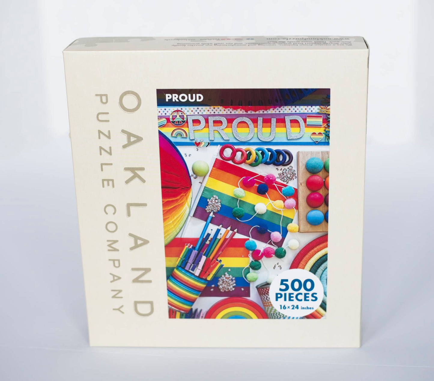 A box branded Oakland Puzzle Company labeled with a photo of colorful image of a variety of objects which are rainbow themed.The word PROUD goes across the top of the image. Overlaying the image is a circle that says 500 Pieces, 16x24 inches