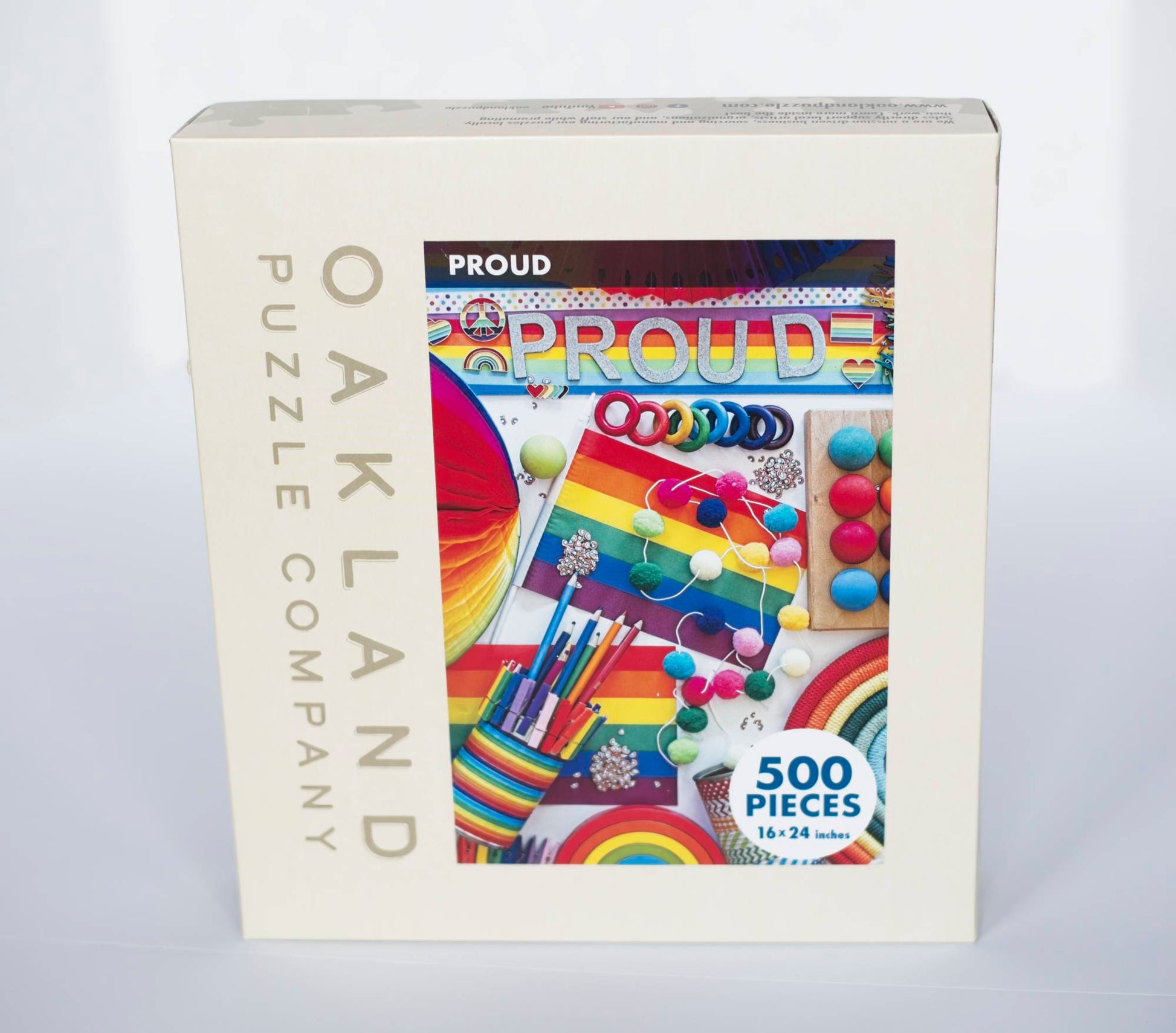 A box branded Oakland Puzzle Company labeled with a photo of colorful image of a variety of objects which are rainbow themed.The word PROUD goes across the top of the image. Overlaying the image is a circle that says 500 Pieces, 16x24 inches