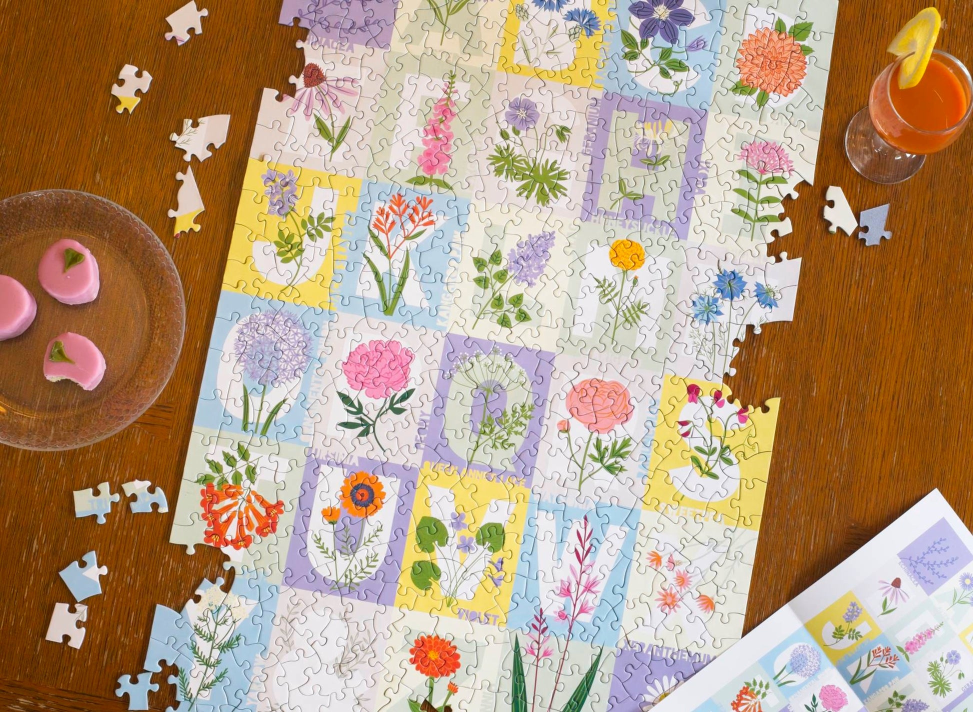 A flatlay shot of the nearly complete ABC FLowers puzzle with just a few pieces scattered around the edges. On the outside of the frame are a glass of carrot juice, the poster for the puzzle, and a plate of small pink cakes, one with a bit taken from it.