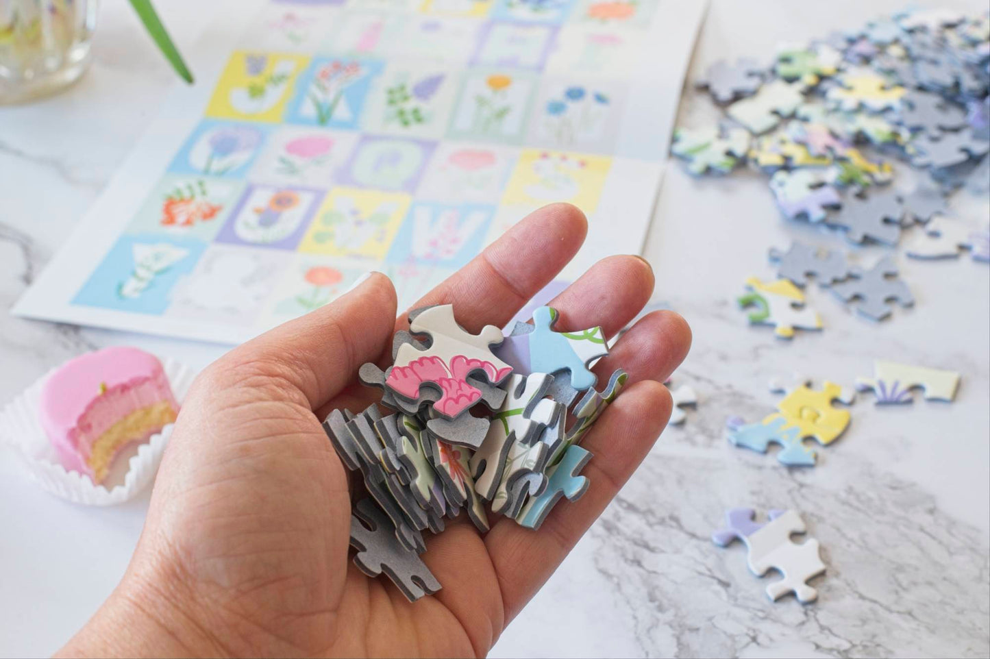 A photo of a hand filled with colorful pieces of the ABC Flowers puzzle with a small pink cake with a bite taken out of it, the poster and other puzzle pieces scattered in the background.