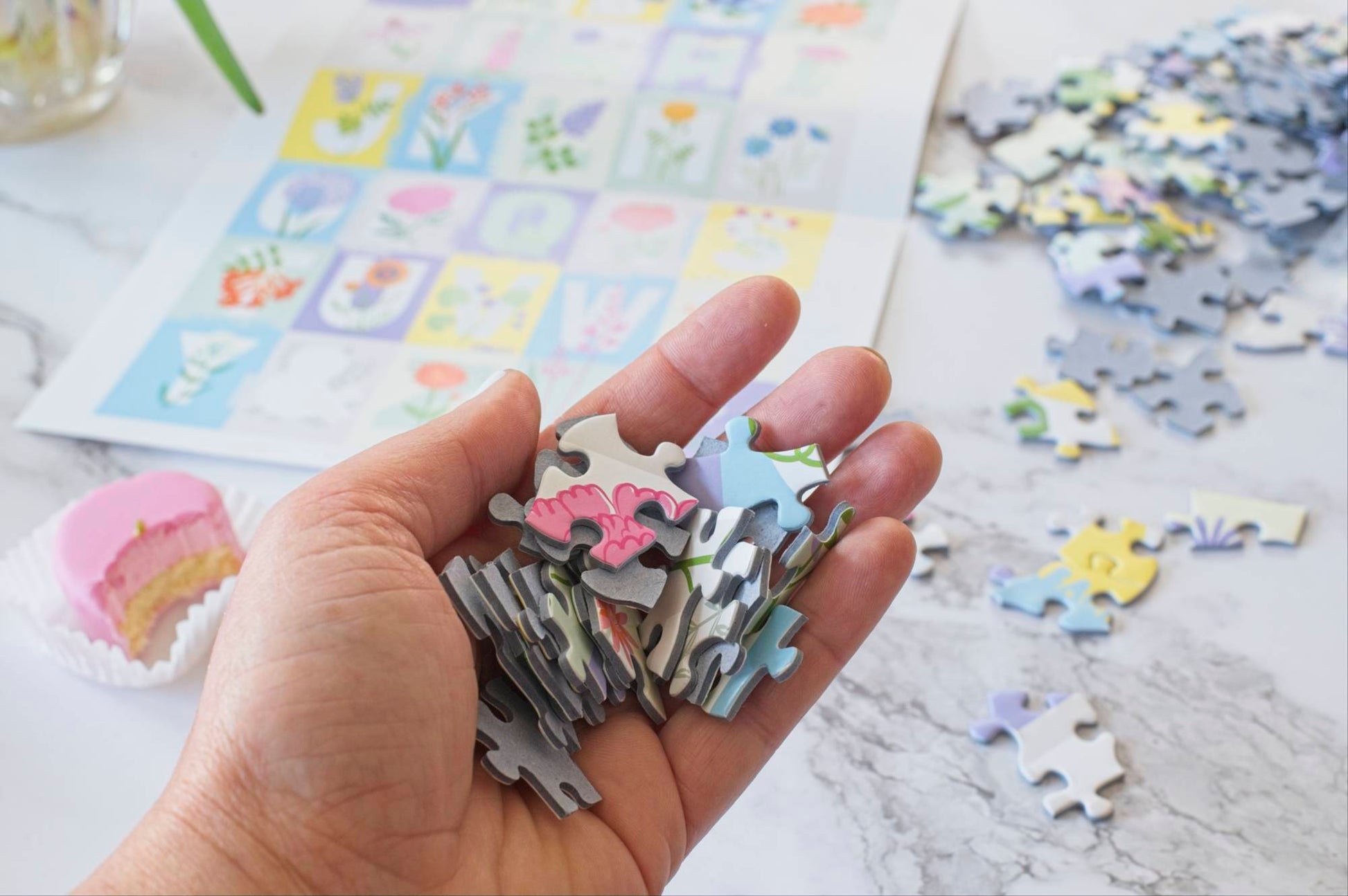 A photo of a hand filled with colorful pieces of the ABC Flowers puzzle with a small pink cake with a bite taken out of it, the poster and other puzzle pieces scattered in the background.