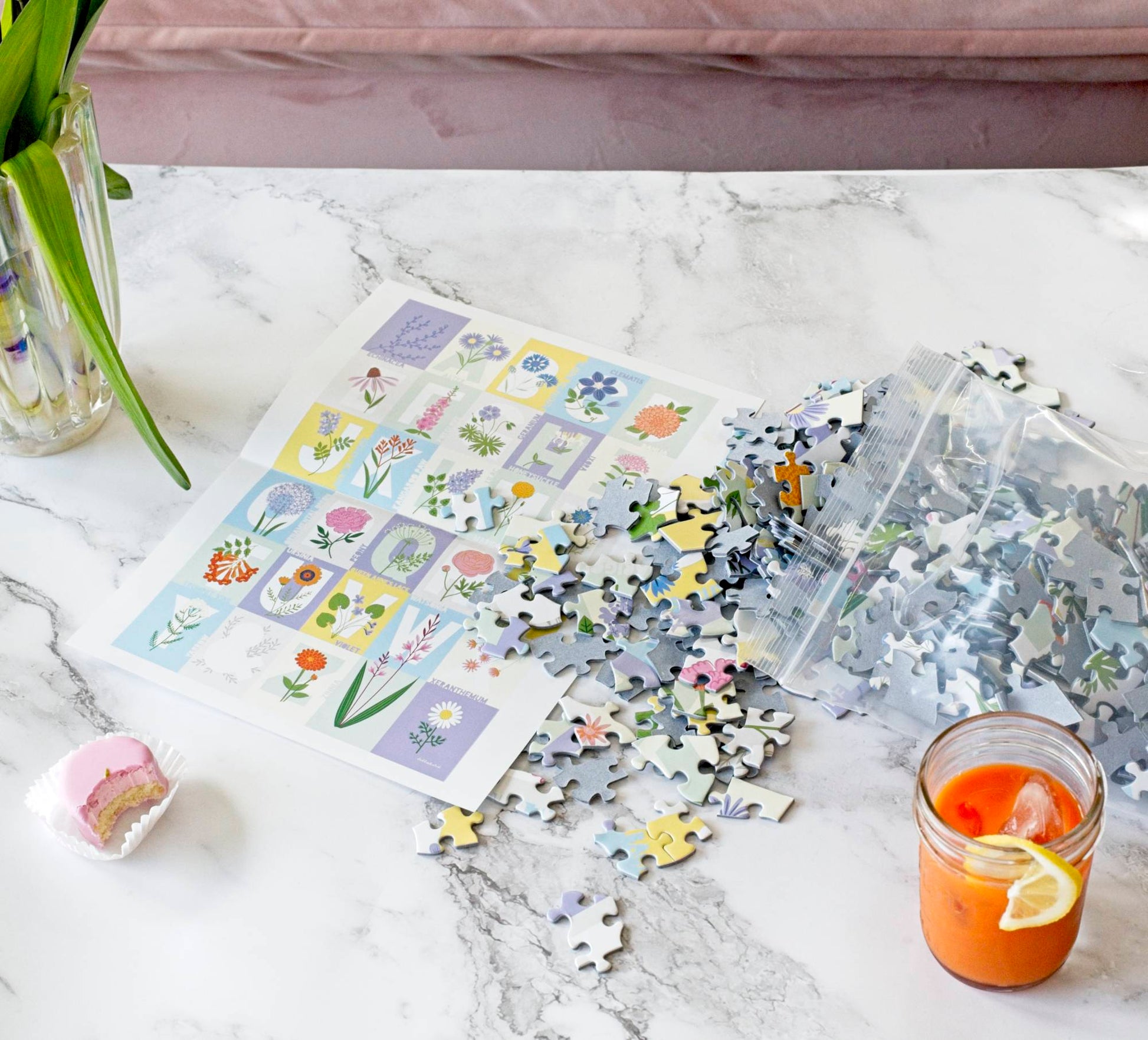 A photograph of a scene with a jar of iced carrot juice, a small piece of cake with a bite out of it and flowers on a marble surface just on the edge of the frame. In the center is a poster of the ABC Flowers puzzle design with a resealable plastic bag spilling pieces out onto it.
