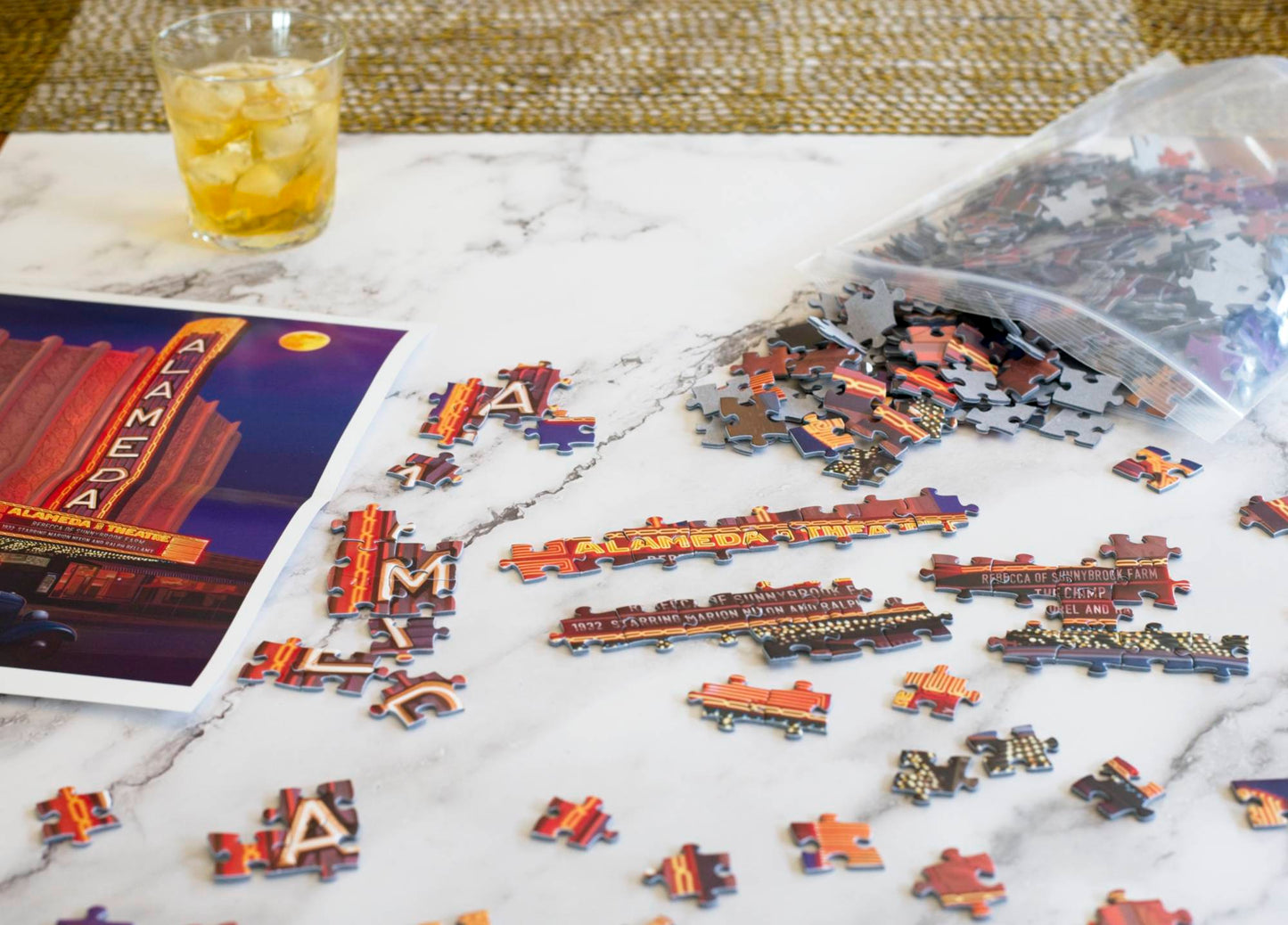 A photo of partially assembled puzzle pieces surrounded by scattered pieces, a poster of the Alameda Theater image, individual pieces spilling from a resealable plastic bag, and a glass of whiskey with ice in the background.