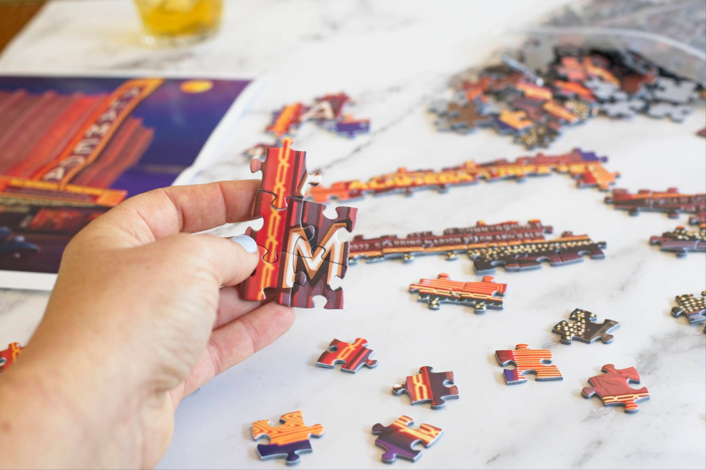 A photo of a hand holding five interlocked jigsaw puzzle pieces of a neon sign showing the letter M, surrounded by partially assembled and scattered puzzle pieces. Blurry in the background, you can see a poster for the Alameda Theater puzzle image and a resealable plastic bag spilling out puzzle pieces.