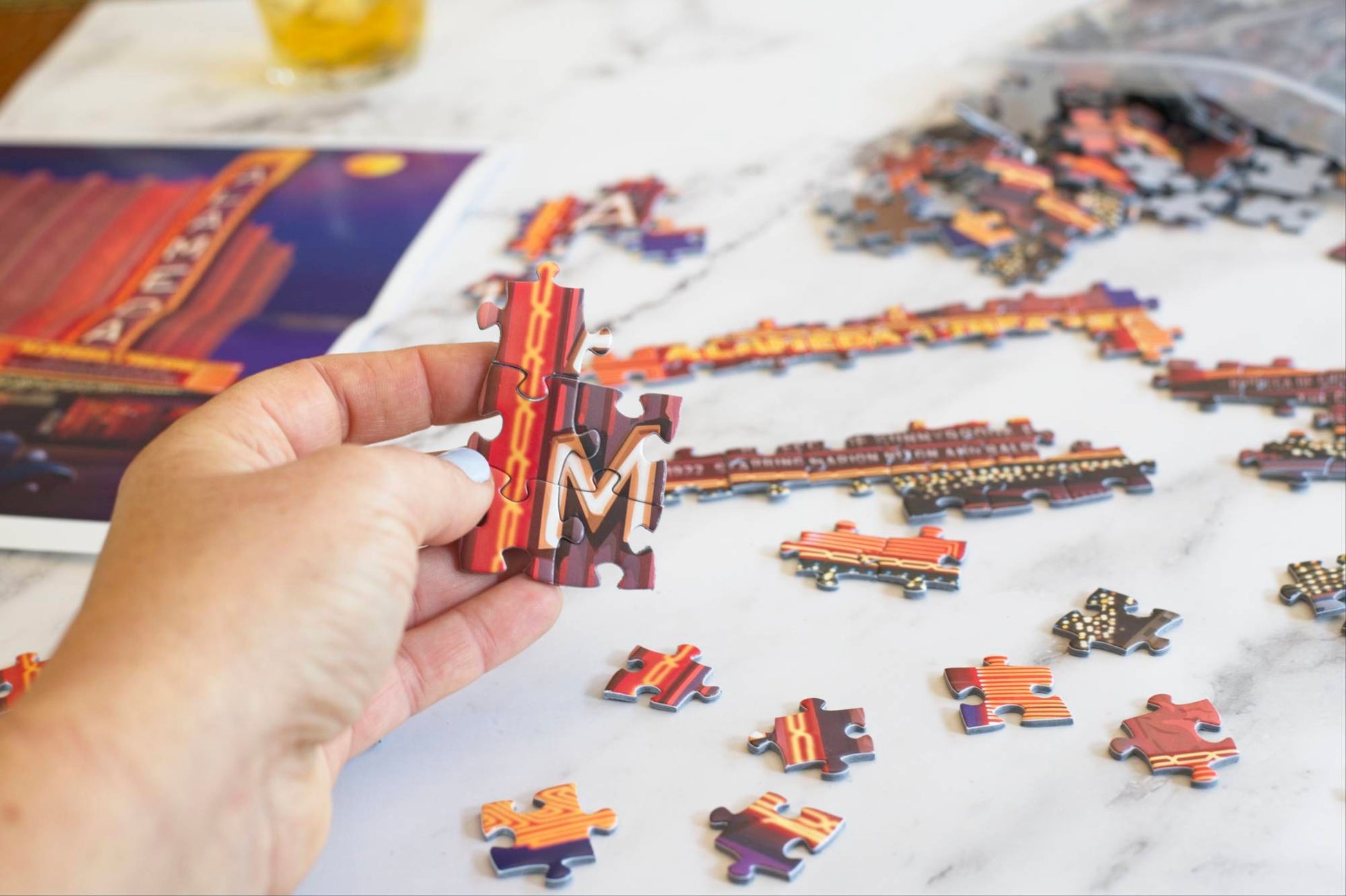 A photo of a hand holding five interlocked jigsaw puzzle pieces of a neon sign showing the letter M, surrounded by partially assembled and scattered puzzle pieces. Blurry in the background, you can see a poster for the Alameda Theater puzzle image and a resealable plastic bag spilling out puzzle pieces.