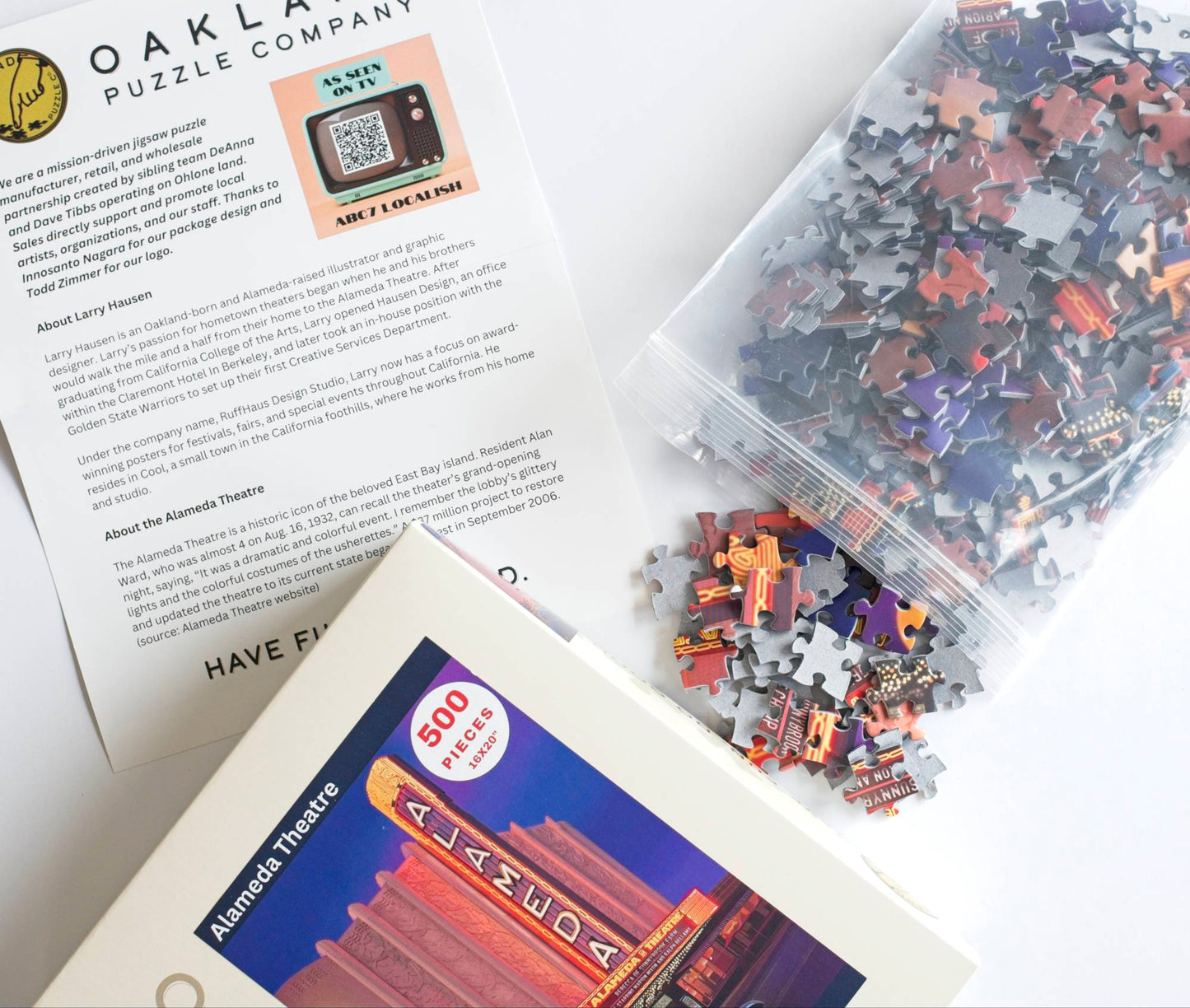 A photo taken at an angle showing a printed description of the Alameda Theater puzzle, artist Larry Hausen, and Oakland Puzzle Company, a product box showing the Alameda Theater image, and a resealable plastic bag with colorful puzzle pieces spilling from it.