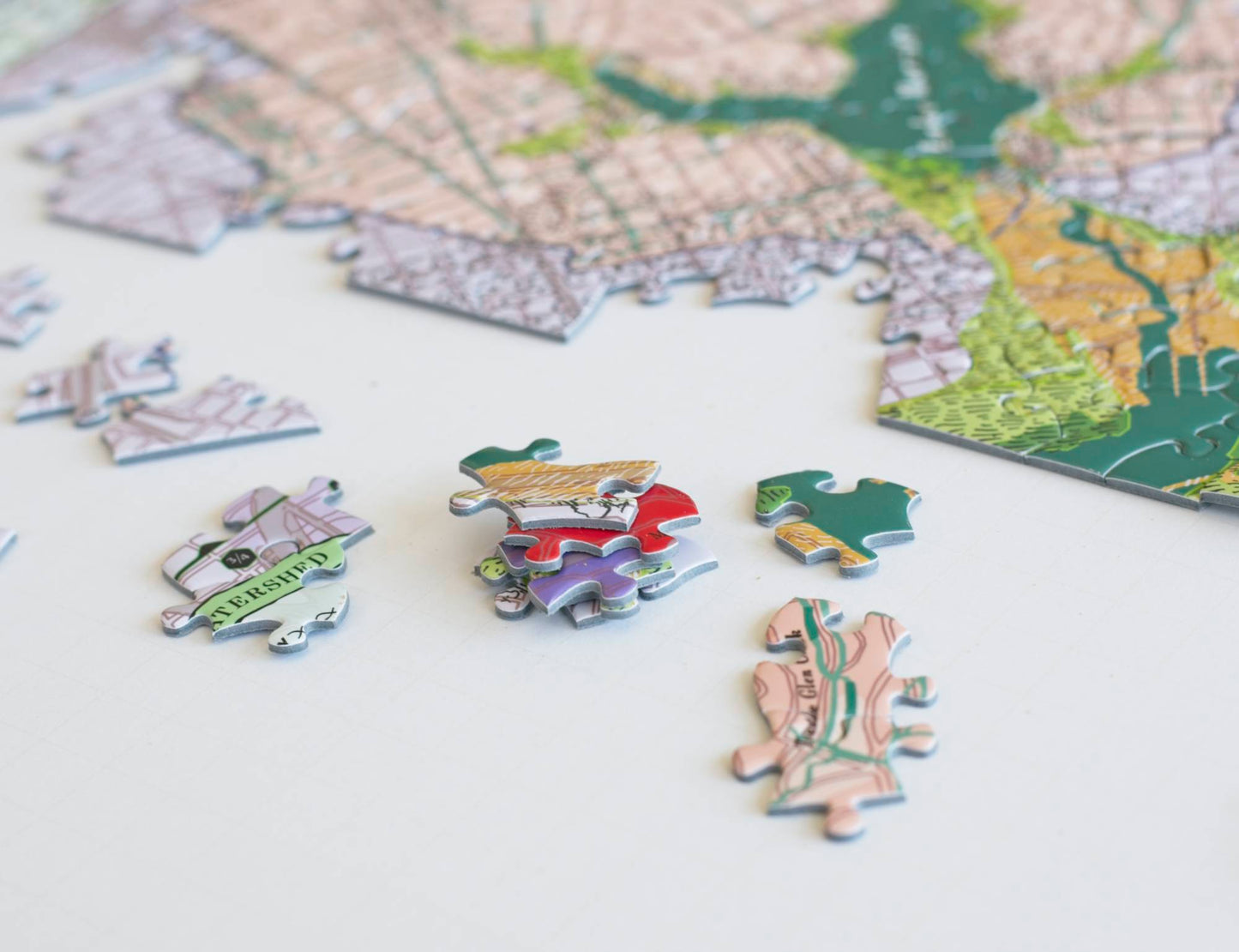 A close up of stacked brightly colored jigsaw puzzle pieces with a few scattered around and a partially completed puzzle blurry in the background