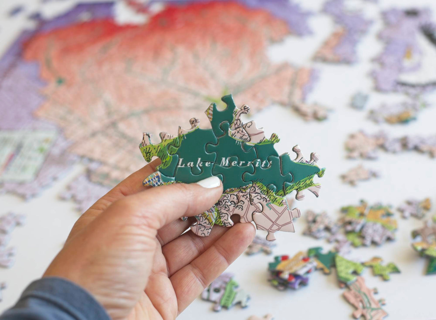 A hand holding a section of a completed jigaw puzzle showing a dark green body of water marked Lake Merritt. Behind the pieces are a partially completed jigsaw puzzle and scattered pieces.