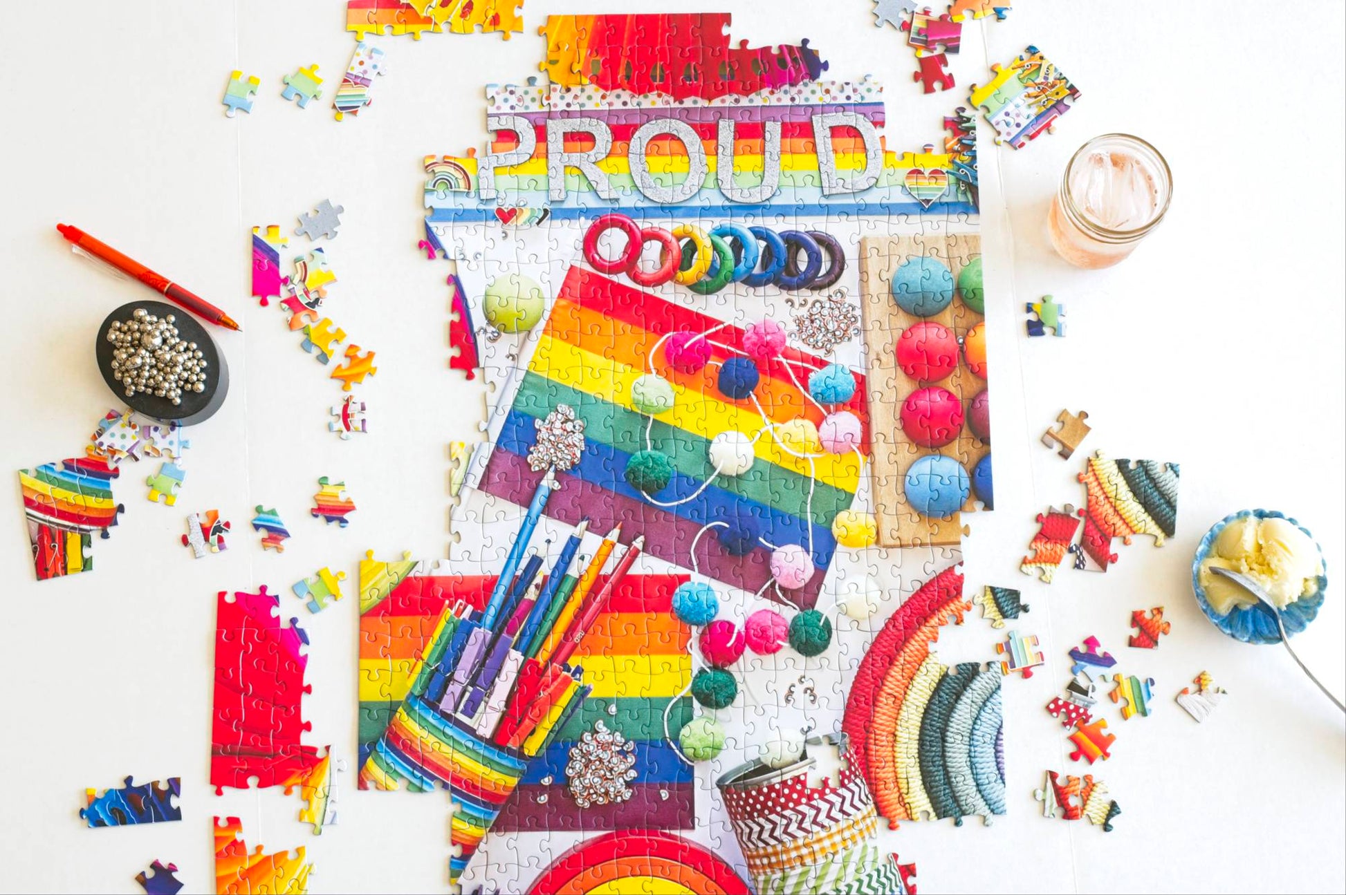 A partially completed rainbow-themed puzzle with a fidget toy, pen, fizzy drink and a bowl of ice cream nearby.
