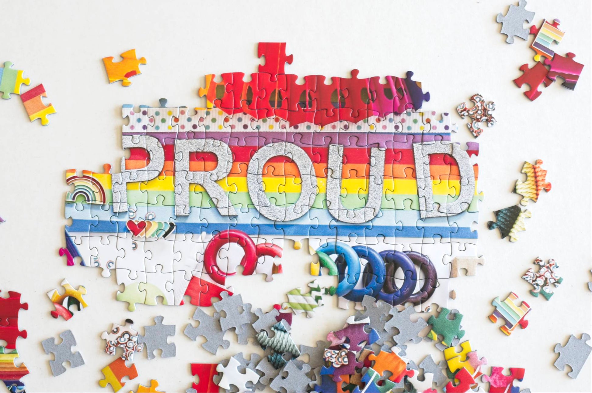 A portion of a puzzle with the word proud in silver glitter on a rainbow colored puzzle sitting on a white background with scattered pieces.
