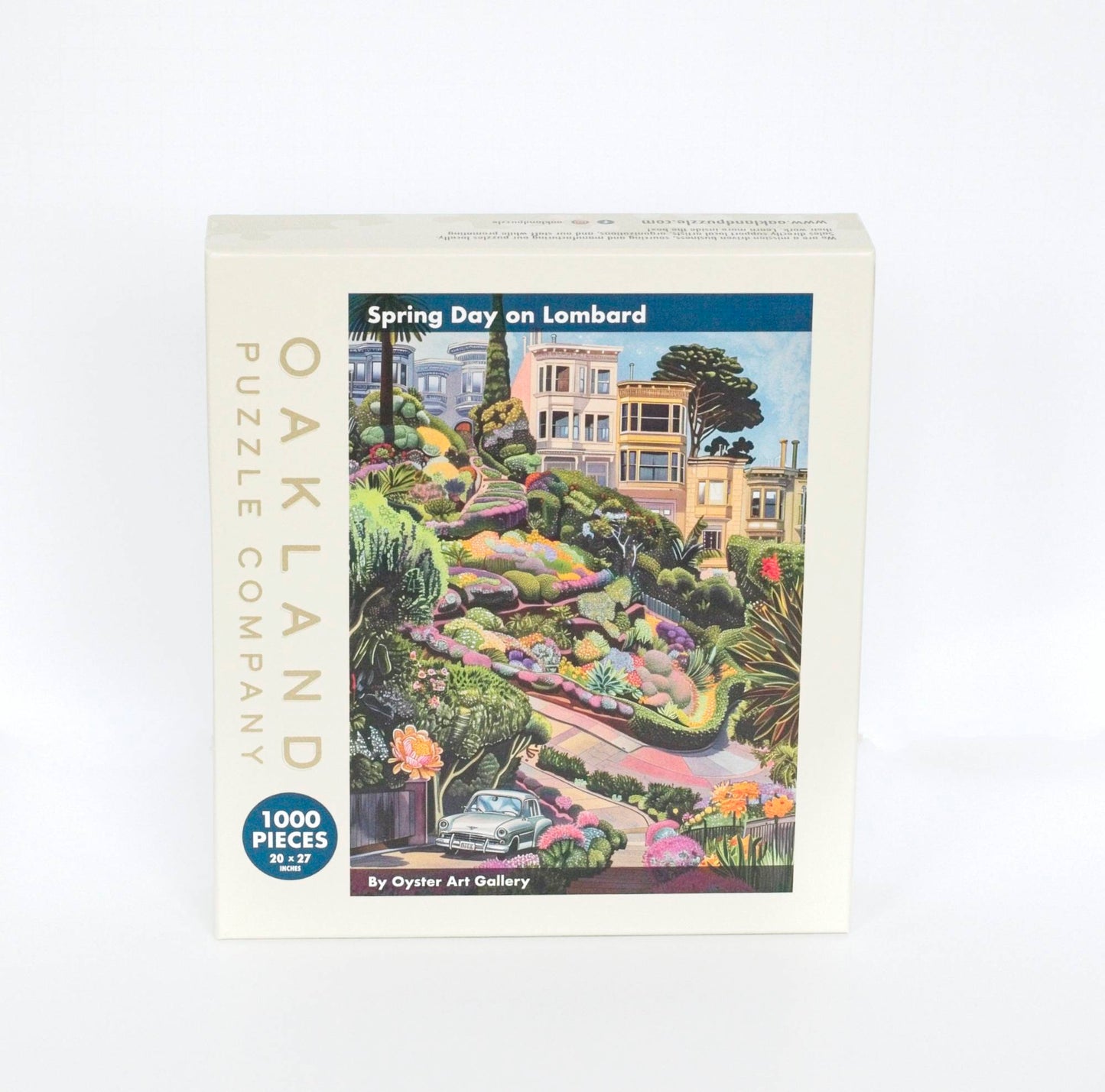Photo of a beige box branded Oakland Puzzle Company. Text Reads Spring Day on Lombard by Oyster Art Gallery 1000 Pieces 20x27 inches. Features a whimsical and colorful image of curvy Lombard Street with Victorian houses behind, a vintage car in the foreground, and a landscape dotted with flower all around.