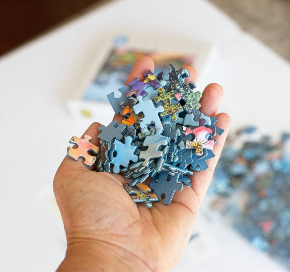 A hand holding a pile of colorful jigsaw puzzle pieces, with a blurred box and more pieces in the background on a white surface.