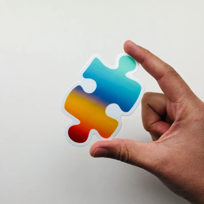 A rainbow colored sticker in the shape of an oversized jigsaw puzzle piece held by a finger and thumb.