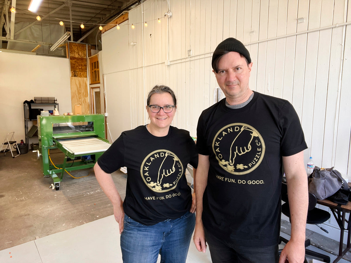 Two people wearing black Oakland Puzzle Company logo t-shirts with gold metallic logo print, standing in front of the die-cut roller press