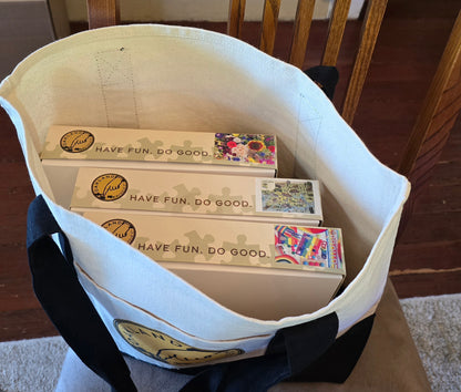 White tote bag with black straps containing puzzle boxes labeled 'Have Fun. Do Good.' on a wooden floor.