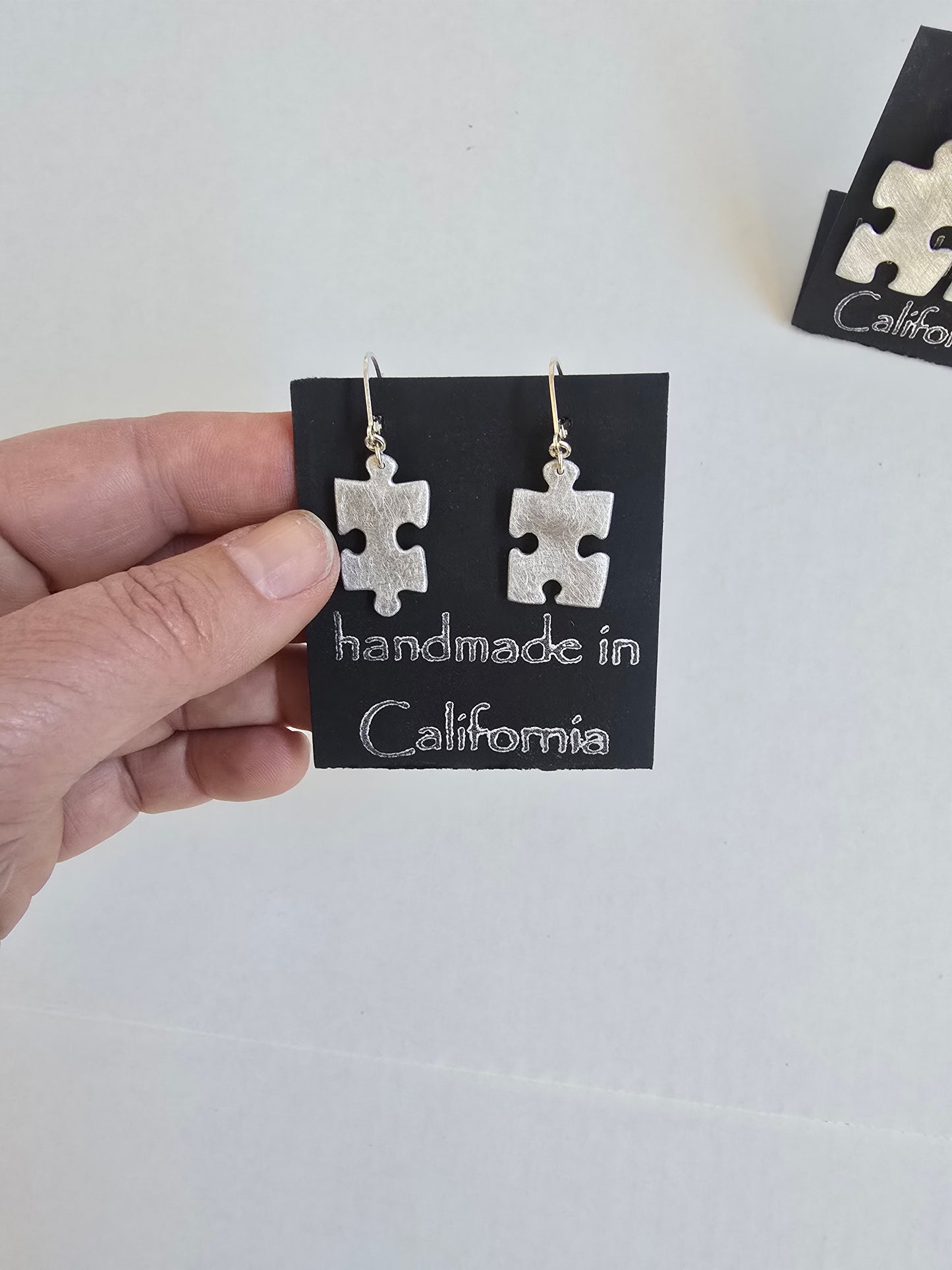 Silver puzzle piece earrings on a 'handmade in California' card held by a hand against a white background.