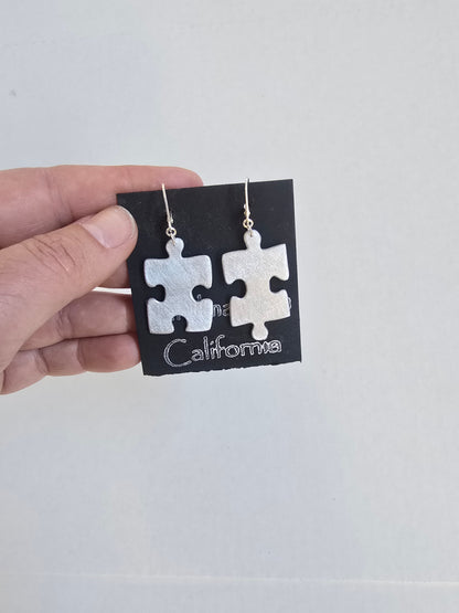 Pair of silver puzzle piece earrings on a black card with 'California' text, held by a hand against a light gray background.