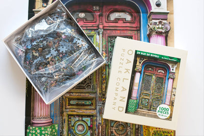 A jigsaw puzzle box open on a reference poster of a colorful doorway. The bottom of the box shows a resealable plastic bag filled with pieces the top has Oakland Puzzle Company.
